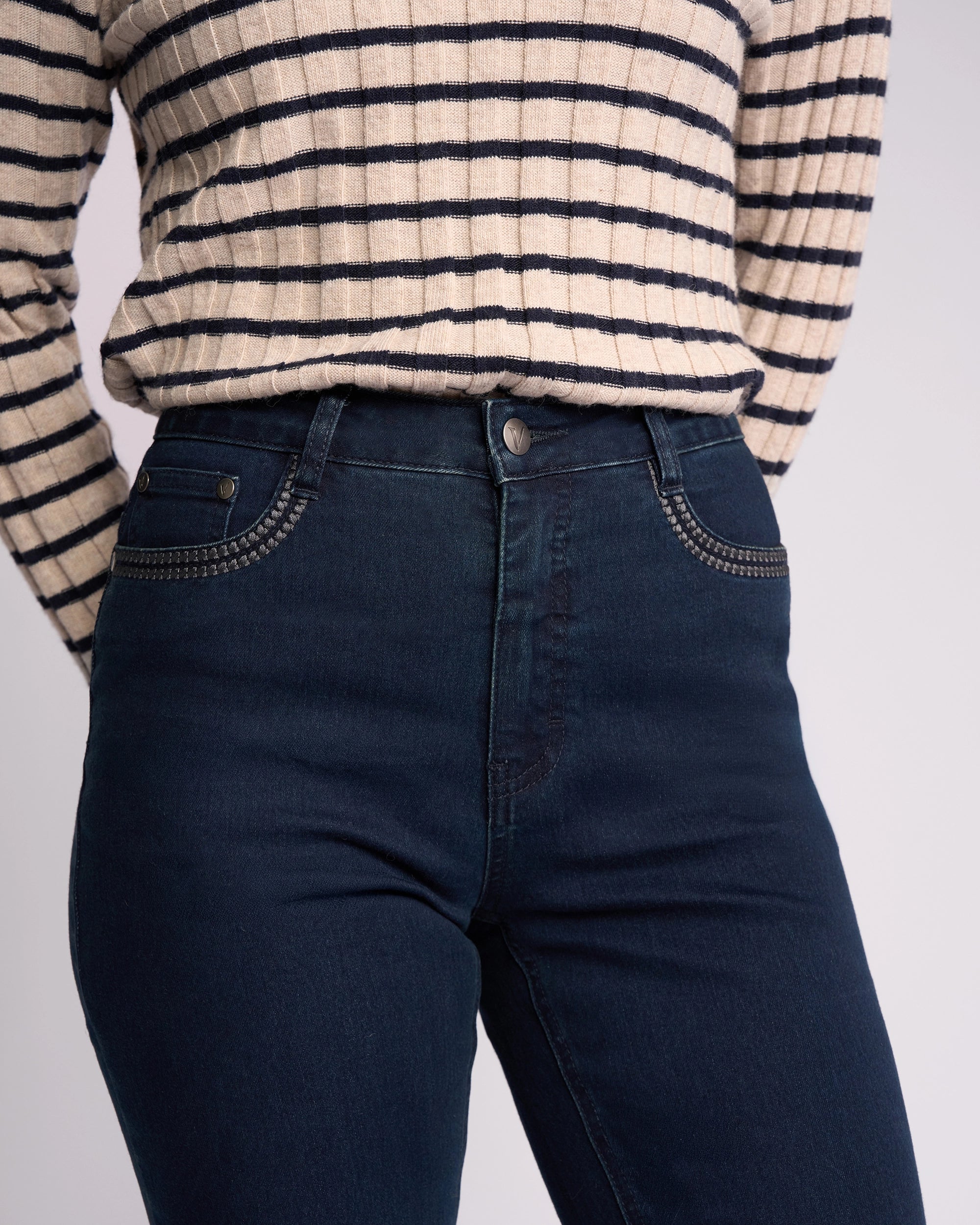 A person models the Vassalli Urban Jean - Mid Blue, styled with a beige sweater featuring black horizontal stripes, tucked in and shown from shoulders to mid-thigh against a plain background.