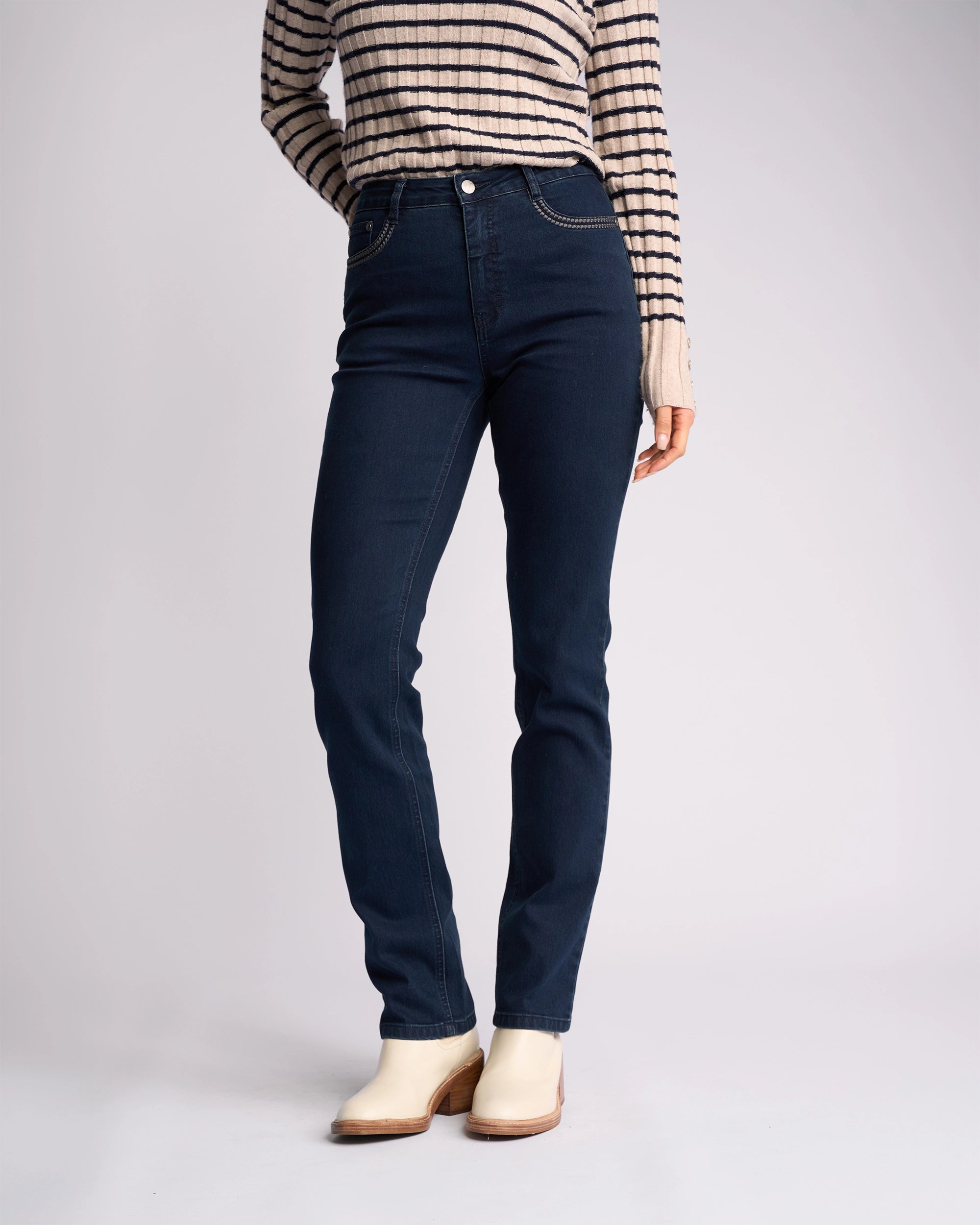 A person wears Vassalli's Urban Jean in Mid Blue, paired with a beige and black striped long-sleeve top and off-white ankle boots, standing against a plain light background. Their head is not visible in the image.