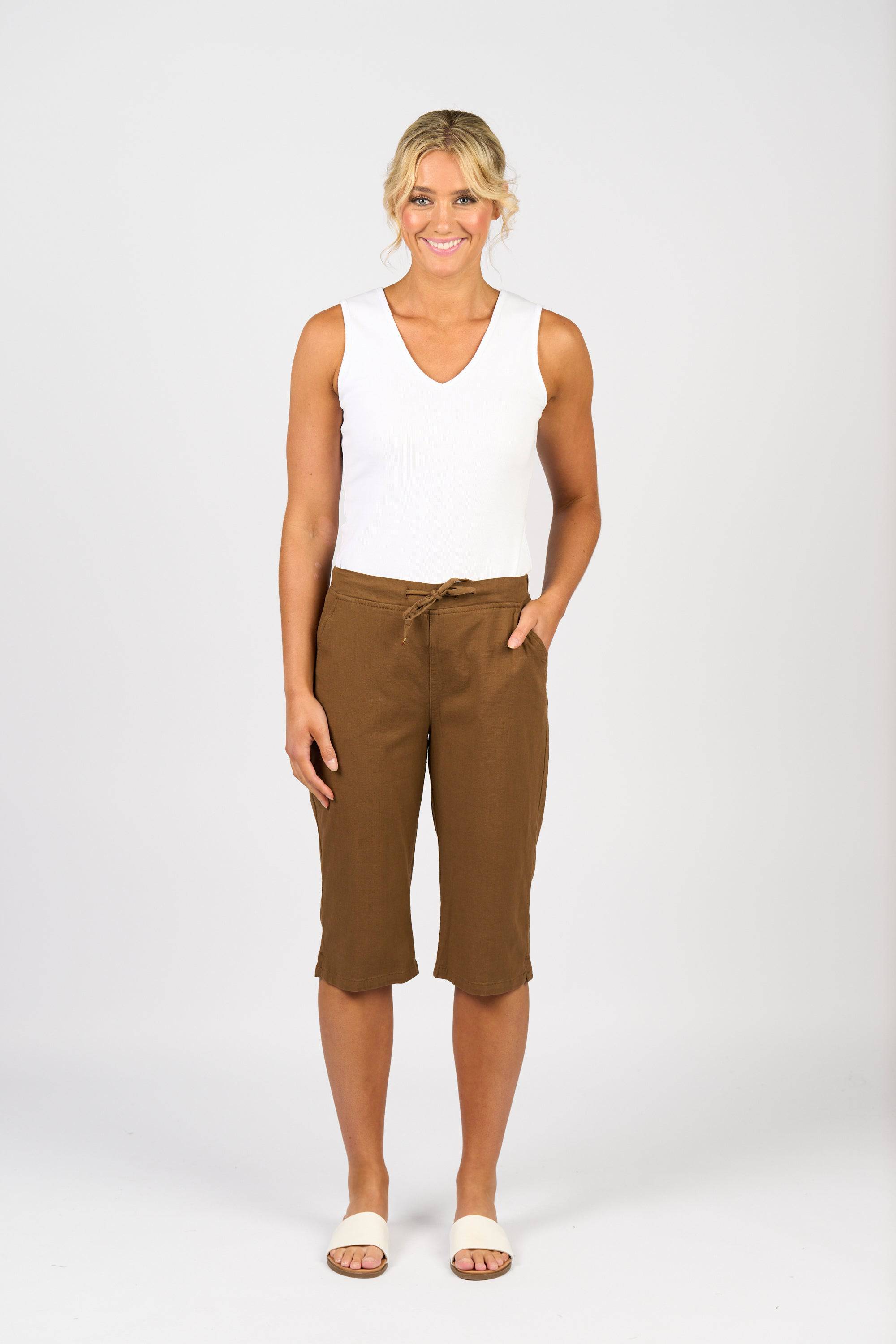 A person stands smiling against a plain white background, wearing a sleeveless white top, Vassalli's Dane Pant in Pecan featuring an elastic waistband, and white slide sandals. One hand is in their pocket.