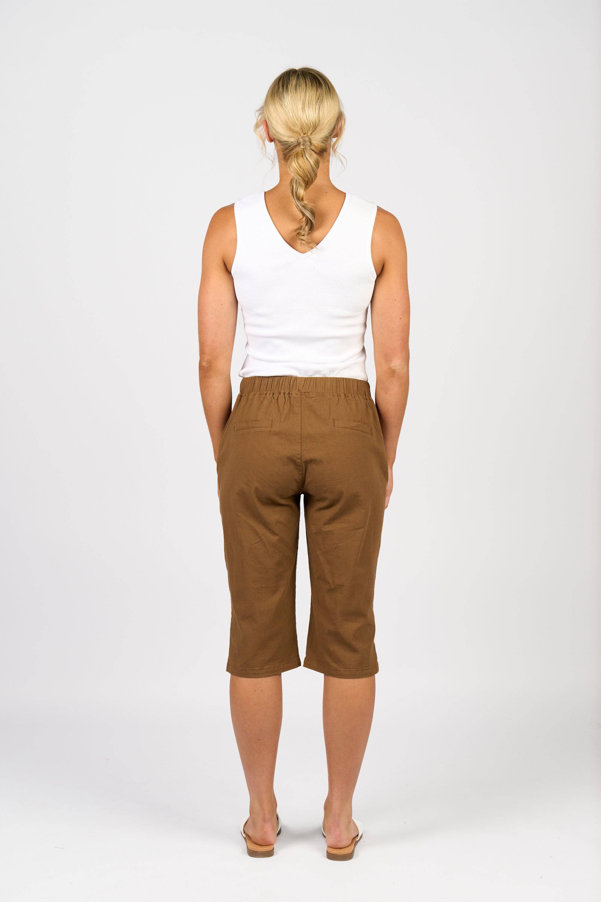 A woman with blonde hair in a loose braid faces away from the camera, wearing a white sleeveless top and Vassalli Dane Pant - Pecan, paired with beige sandals. The stretch cotton-viscose pants are knee-length with an elastic waistband. Background is plain white.