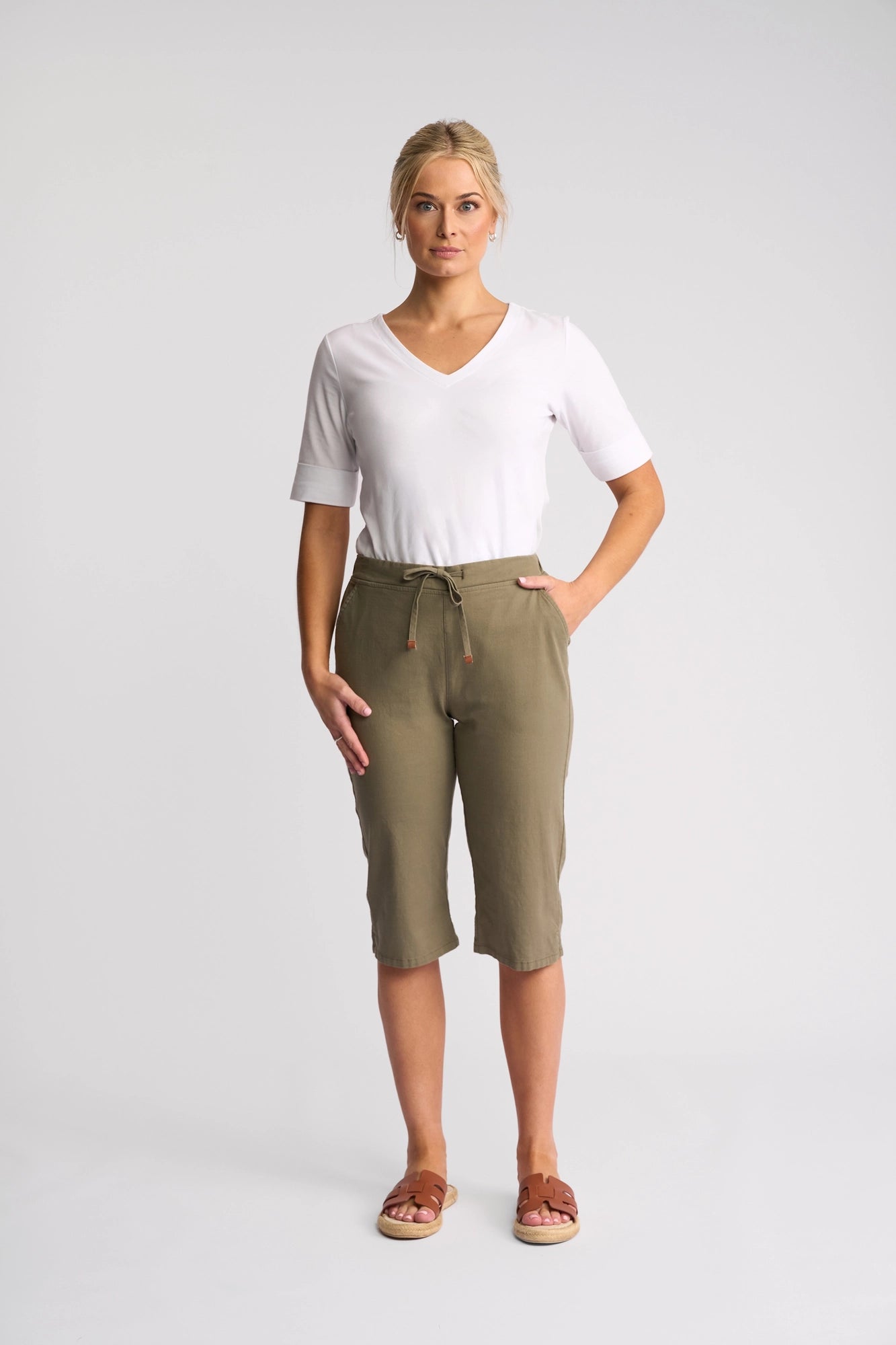 A woman stands against a plain background, wearing the Vassalli Drew 3/4 Pant in Army—olive green, knee-length pants crafted from stretch cotton with a comfortable pull-on elastic waistband. She pairs them with a white V-neck T-shirt and relaxed brown sandals, her hands casually tucked in her pockets as she faces forward.
