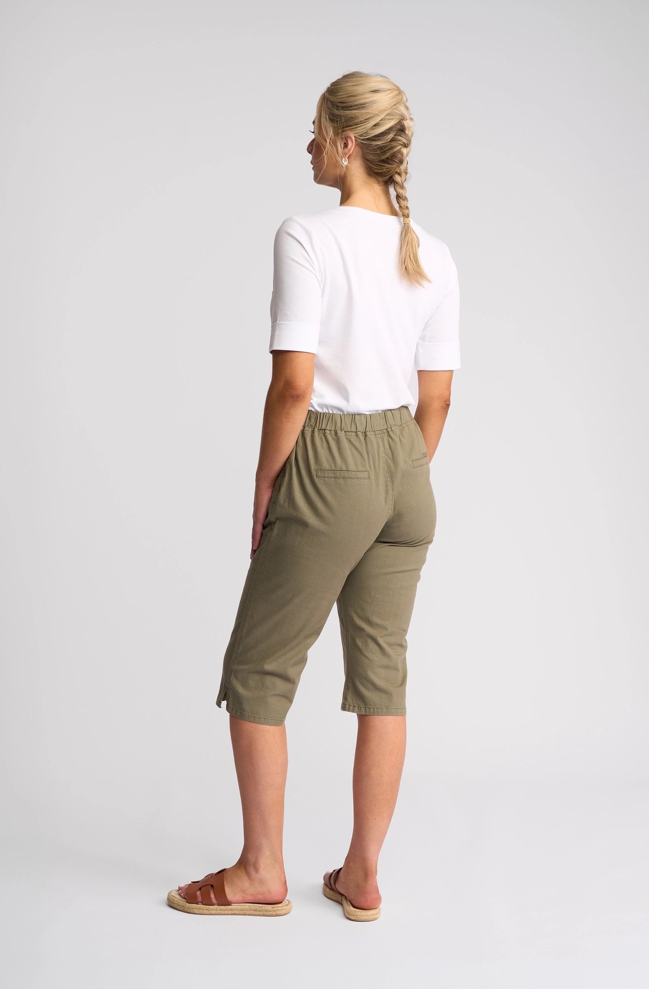 A person with blonde hair in a braid is standing, facing away. They wear a white top, Vassalli Drew 3/4 Pant in Army—an olive green slim-leg pant with a pull-on elastic waistband made from stretch cotton—and brown sandals against a plain light background.
