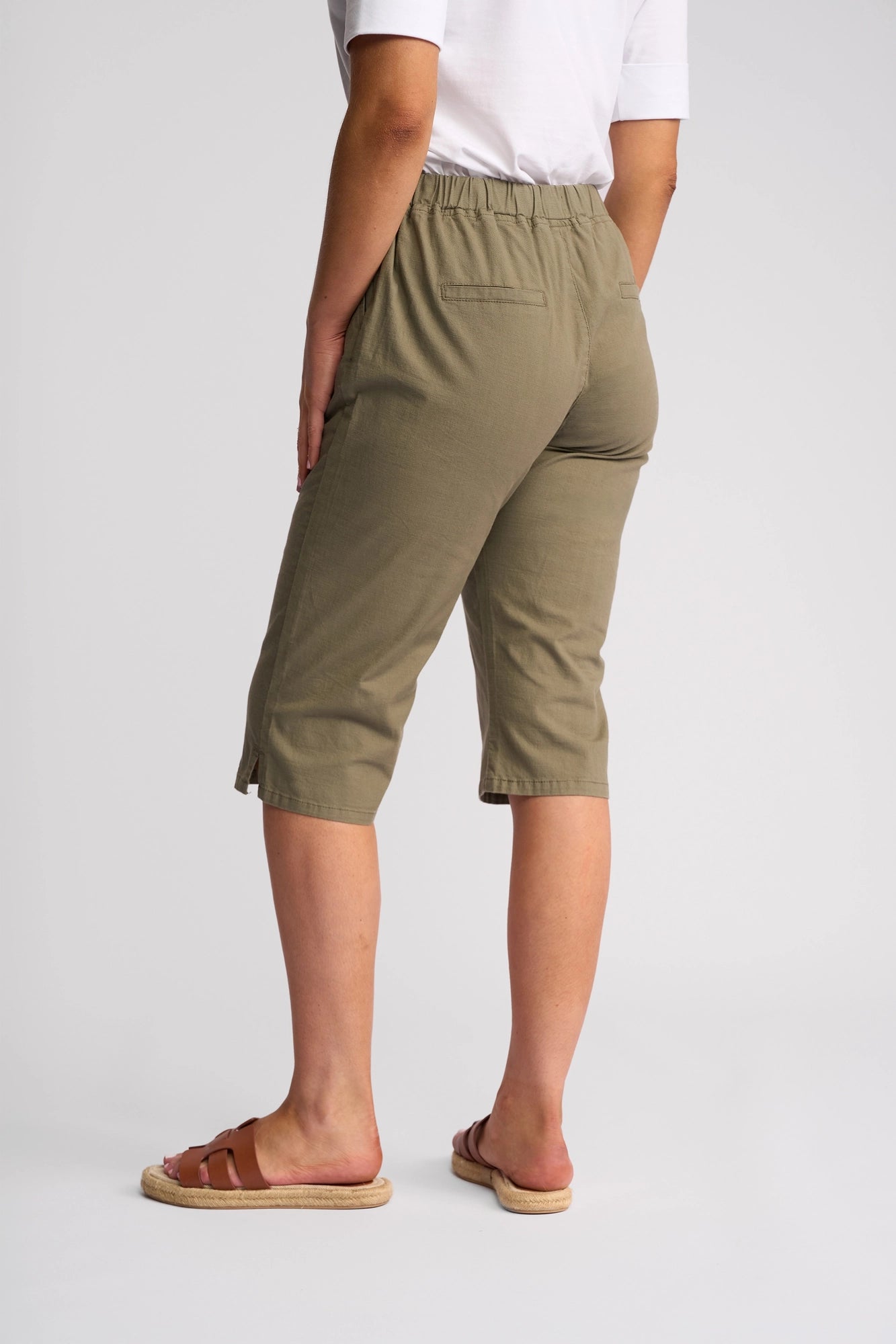A person stands facing away against a plain white background, wearing a white shirt and the Vassalli Drew 3/4 Pant in Army—olive green, knee-length pants with a pull-on elastic waistband—paired with brown sandals and arms relaxed by their sides.