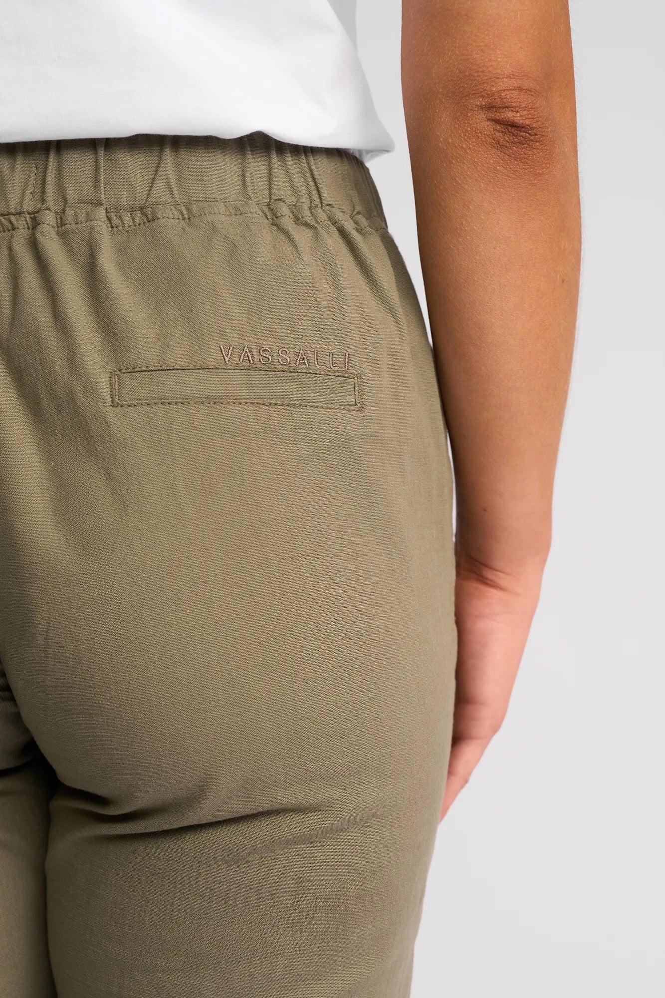 A close-up of a person wearing the Vassalli Drew 3/4 Pant in Army, featuring an olive green slim-leg design crafted from stretch cotton with a comfortable pull-on elastic waistband. “Vassalli” is embroidered above the back pocket, and the person is standing against a plain, light background.