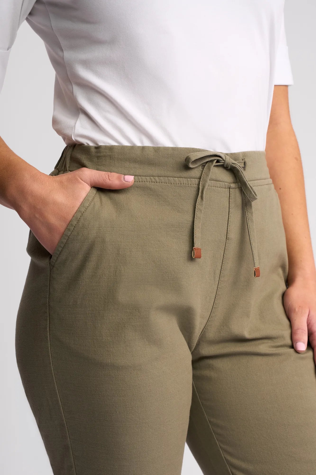 A person is seen wearing the Vassalli Drew 3/4 Pant in Army, featuring a pull-on elastic waistband, drawstring detail, and practical pockets. These slim-leg pants are crafted from stretch cotton and are paired with a white top. The image is cropped from mid-torso to mid-thigh, with one hand casually resting in a pocket.