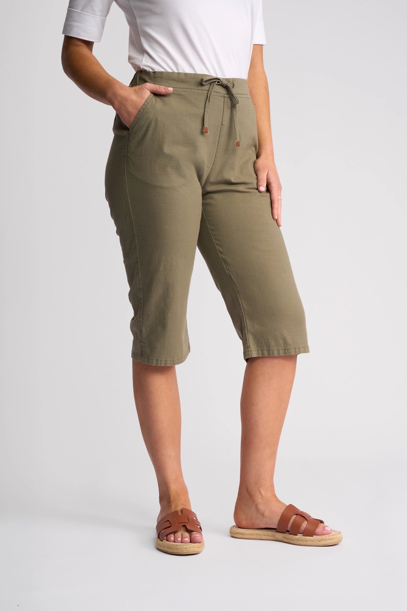 A person wearing the Vassalli Drew 3/4 Pant in Army, styled with a white short-sleeve top and brown slide sandals, stands with one hand in their pocket against a plain light background.