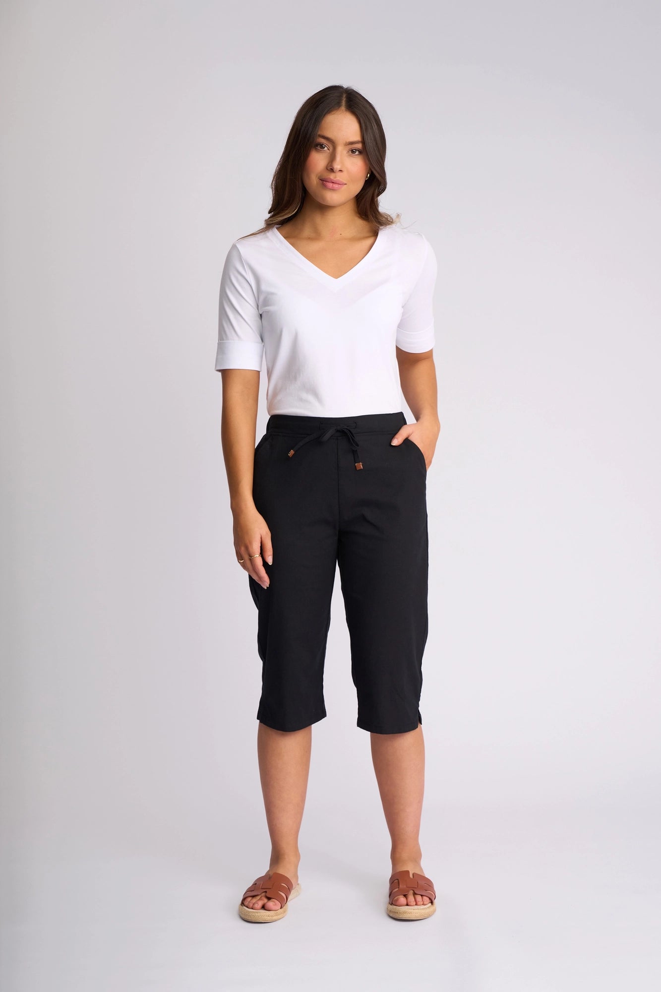 A woman stands facing forward, wearing a white V-neck shirt and the Vassalli Drew 3/4 Pant in Black. The slim-leg pants are made from stretch cotton and are paired with brown sandals. She has one hand in her pocket and a neutral expression, set against a plain light grey background.