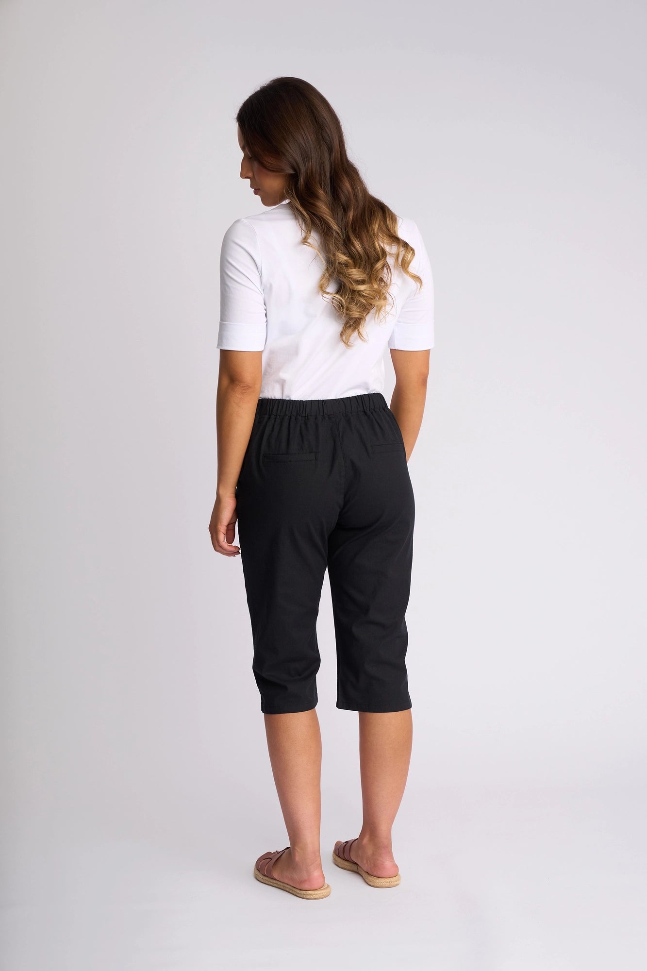 A woman with long wavy hair, dressed in a white short-sleeve top, Vassalli Drew 3/4 Pant in black with a stretch cotton slim-leg fit, and sandals, stands facing away against a plain white background.