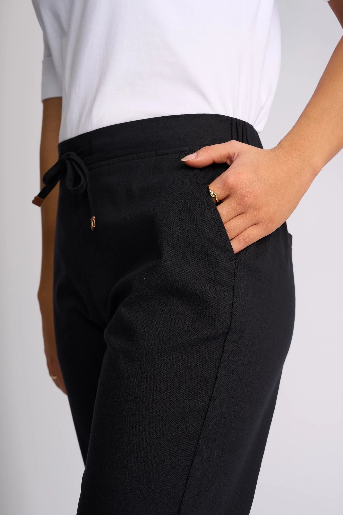 A person wearing the Vassalli Drew 3/4 Pant in black—a slim-leg style made from stretch cotton—and a white top places their right hand in the pant’s pocket. The image is cropped to display only the torso and upper legs.