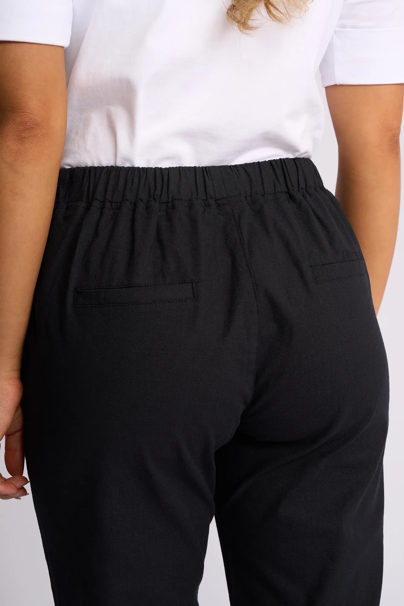 Model is seen from the back and waist down, wearing a white shirt paired with the Vassalli Drew 3/4 Pant in Black. The slim-leg pant features an elastic waistband and two back welt pockets. Part of one arm and hand is also visible.