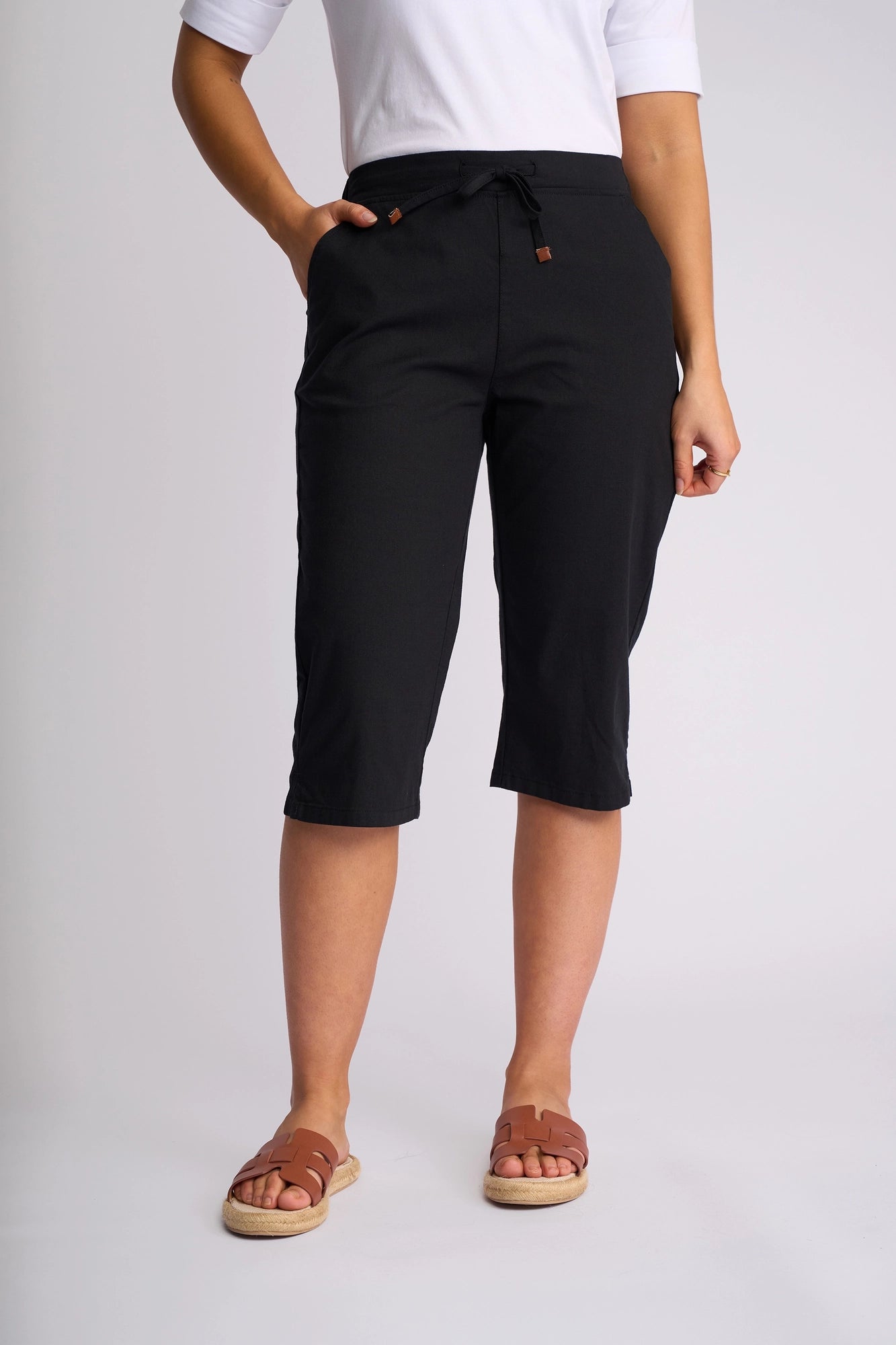 A person is shown from the waist down, wearing a white shirt paired with Vassalli Drew 3/4 Pant in black—slim-leg pants with pockets, crafted from stretch cotton—and brown sandals, against a plain white background.