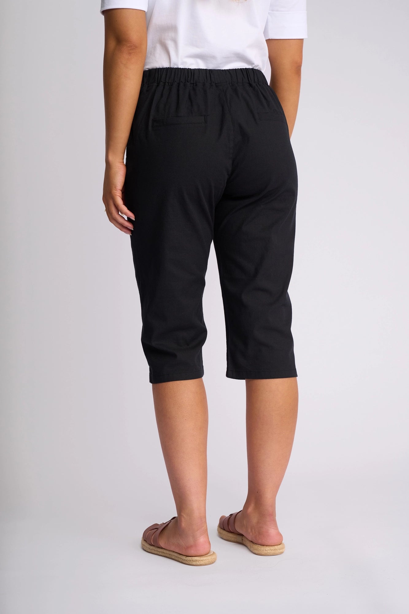 A person wearing a white shirt, the Vassalli Drew 3/4 Pant in black, and sandals stands facing away from the camera against a plain background.