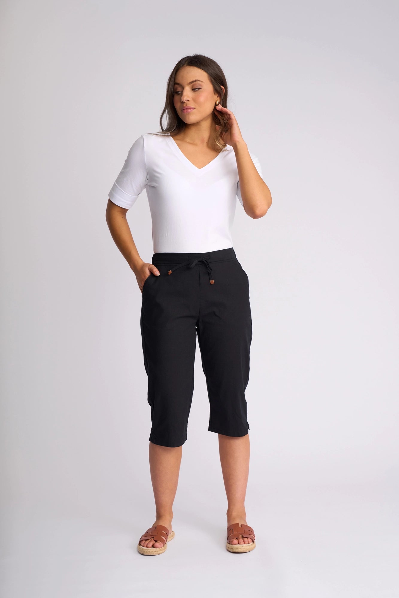 A woman stands against a plain background wearing a white V-neck top and the Vassalli Drew 3/4 Pant in Black—crafted from stretch cotton for comfort and featuring a slim-leg silhouette—paired with brown sandals. She has one hand in her pocket and the other touching her hair.