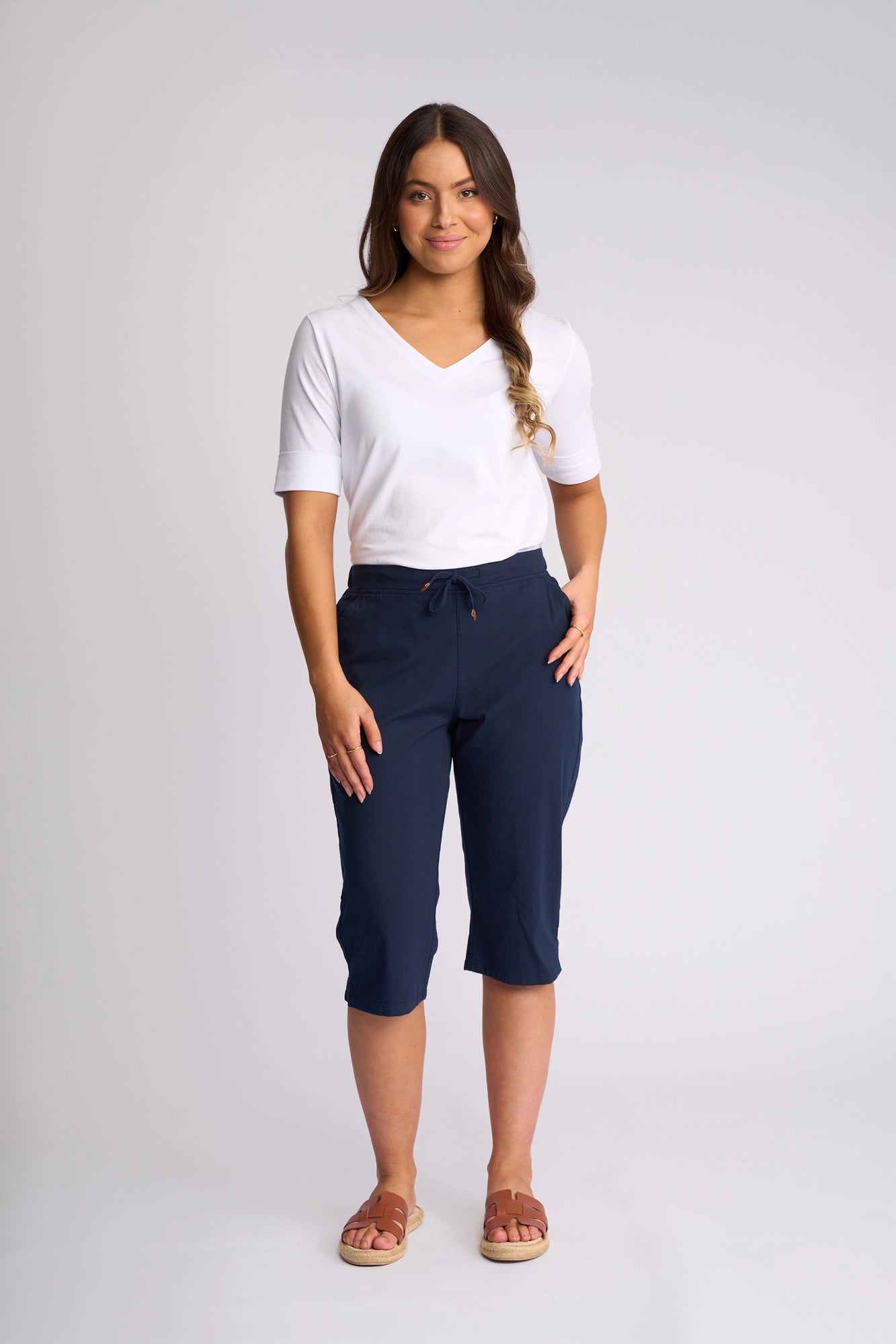 A woman stands against a plain background, smiling with one hand on her hip, wearing a white short-sleeve V-neck top, Vassalli Drew 3/4 Pant in Ink—crafted from comfortable stretch cotton with a drawstring—and sandals.