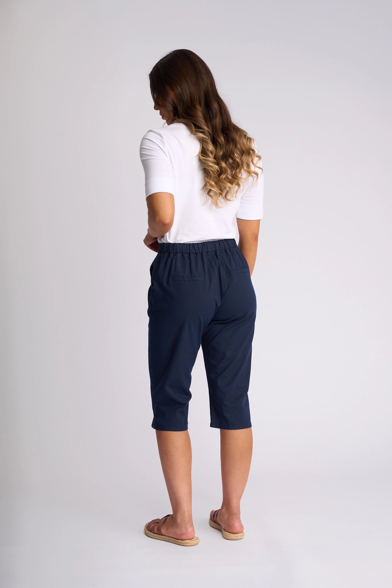 A woman stands facing away, wearing a white short-sleeve top and Vassalli Drew 3/4 Pant in Ink—a slim-leg pant crafted from stretch cotton—paired with sandals. Her long wavy hair cascades down her back against a plain light background.