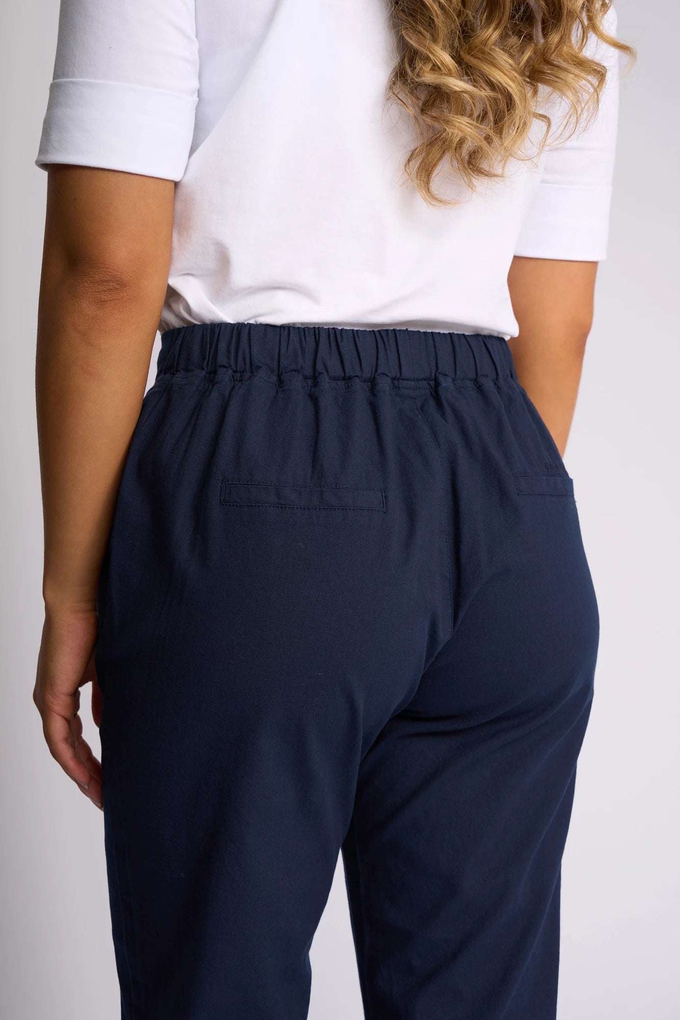 A person is shown from the back wearing the Vassalli Drew 3/4 Pant in Ink, featuring a slim-leg silhouette and elastic waistband, paired with a white short-sleeve shirt. Their wavy hair falls over one shoulder.