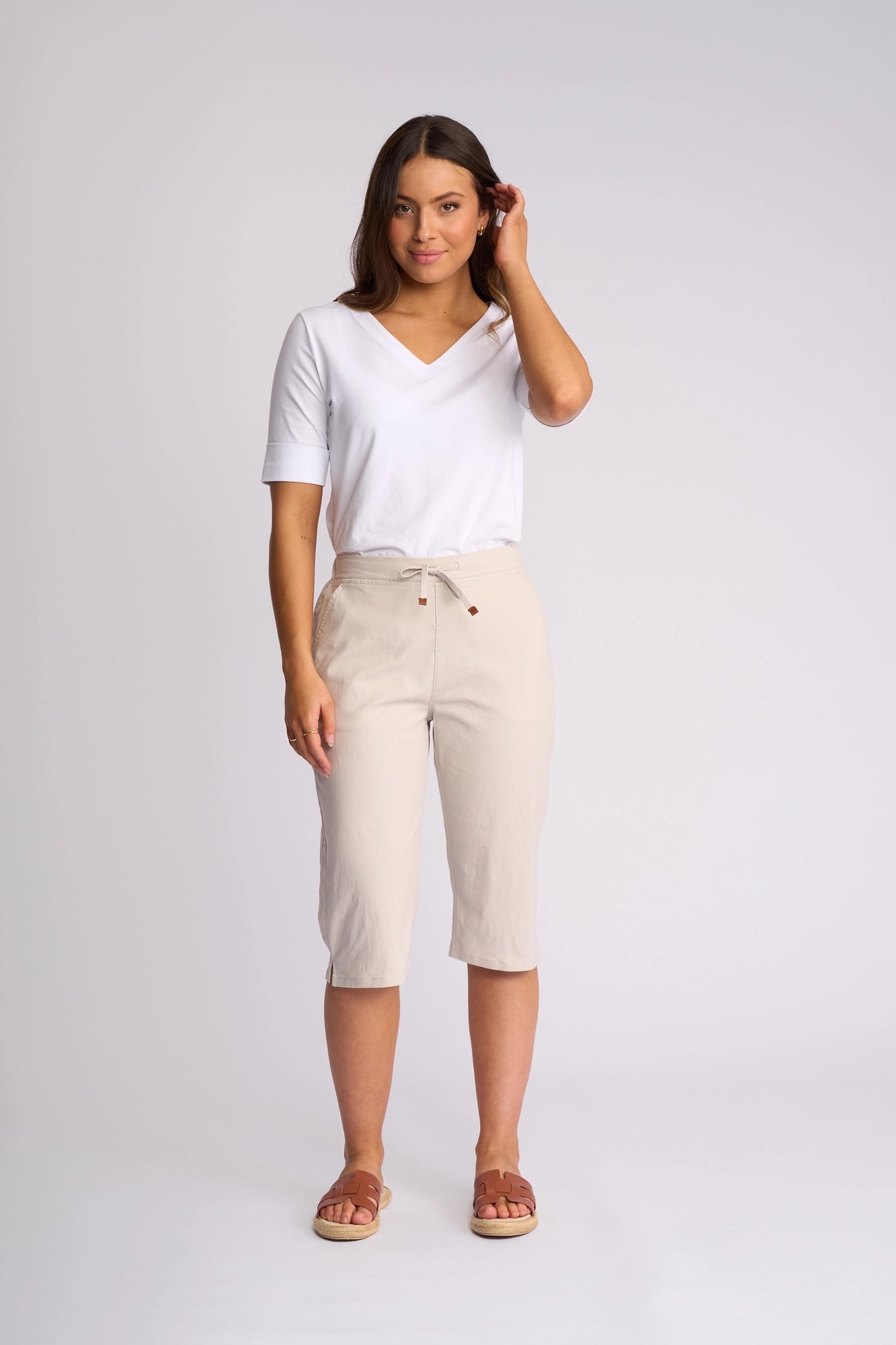 A woman stands against a plain background wearing the Vassalli Drew 3/4 Pant in Stone—a light beige, slim-leg style with a pull-on elastic waistband in soft stretch cotton—paired with a white V-neck top and brown sandals. She has long brown hair and touches her hair with one hand.
