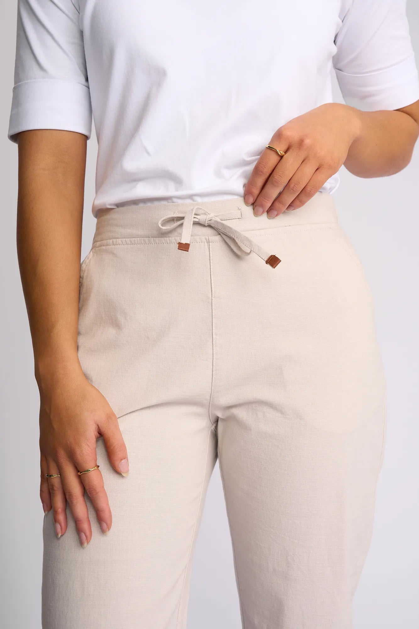 A person wearing a white short-sleeved shirt and Vassalli’s Drew 3/4 Pant in Stone, featuring a pull-on elastic waistband and slim-leg stretch cotton design. One hand rests on the waistband while the other is by their side, with only the torso and upper legs visible to highlight the comfortable fit of these pants.