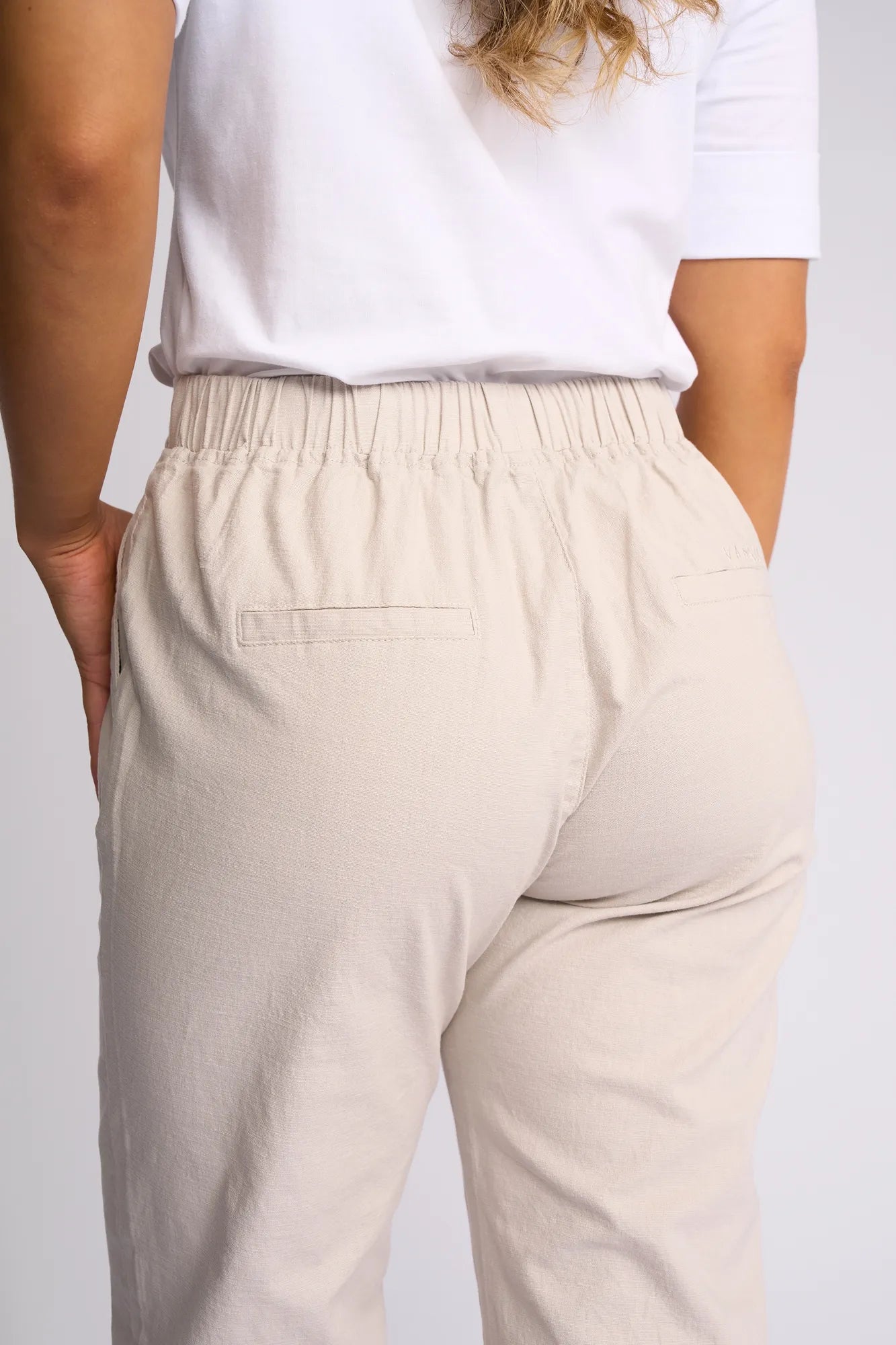 A person is shown from the back wearing the Vassalli Drew 3/4 Pant in Stone—a light beige, slim-leg pant featuring a pull-on elastic waistband and two back welt pockets for added ease. Crafted from stretch cotton fabric for all-day comfort, the look is completed with a white short-sleeve shirt, as the person’s left arm rests by their side.