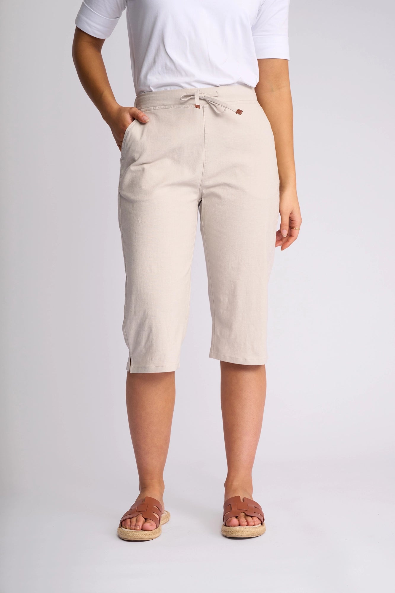 A person wearing a white t-shirt, the Vassalli Drew 3/4 Pant in Stone—beige knee-length drawstring shorts crafted from stretch cotton—and brown slide sandals stands against a plain light background.