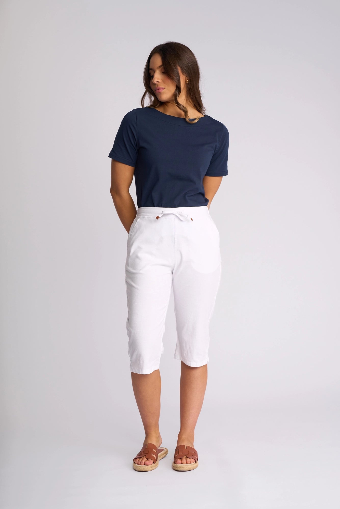 A woman stands against a plain background, wearing a navy blue t-shirt and Vassalli Drew 3/4 Pant in white—crafted from stretch cotton—paired with brown sandals. She has her hands behind her back and is looking to the side.