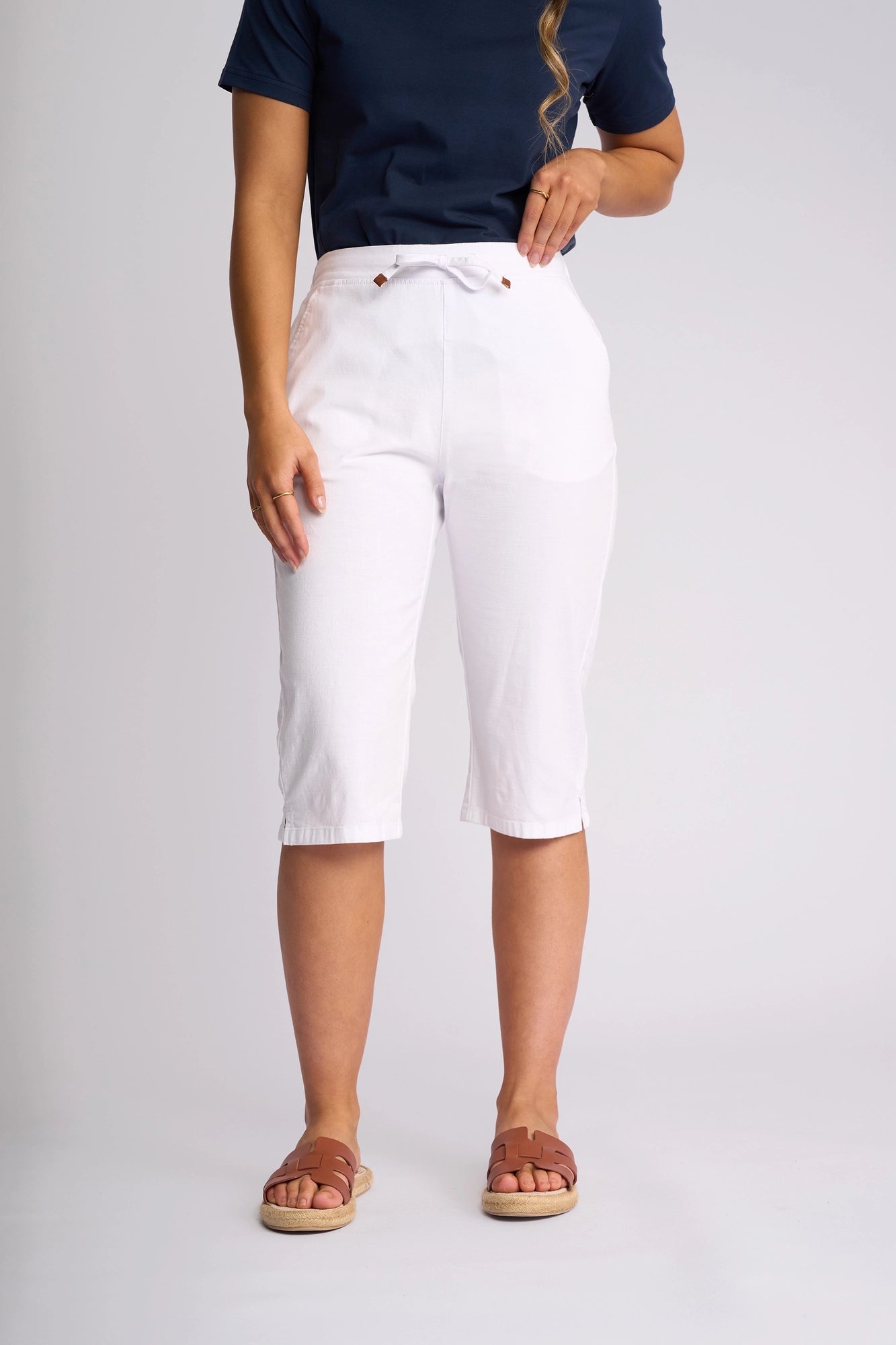 A person stands against a plain light background wearing a navy blue shirt, brown sandals, and the Vassalli Drew 3/4 Pant in white—a slim-leg style made from stretch cotton with a drawstring. Their face is not visible.