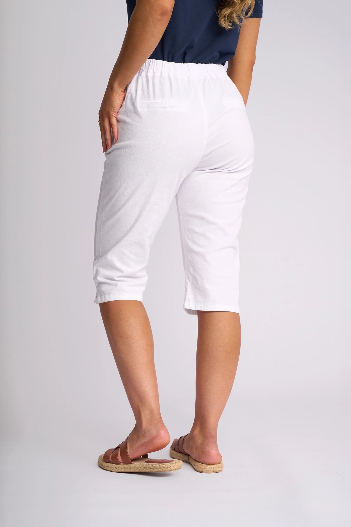 A person stands facing away, wearing the Vassalli Drew 3/4 Pant in White—crafted from stretch cotton with a slim-leg silhouette—paired with a navy blue top and pink sandals. The background is plain light gray.