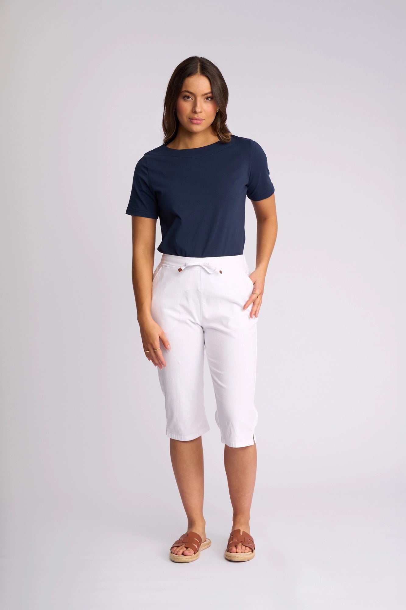 A woman stands against a plain background wearing the Vassalli Drew 3/4 Pant in white, paired with a navy blue t-shirt and brown sandals. She has one hand on her hip and a neutral expression.
