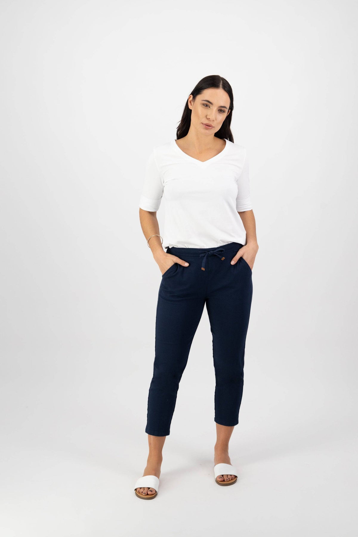 A woman with long dark hair wears a white V-neck top, Vassalli Denver 7/8 Pant in Ink, and white slide sandals, standing against a plain white background with her hands in her pockets.
