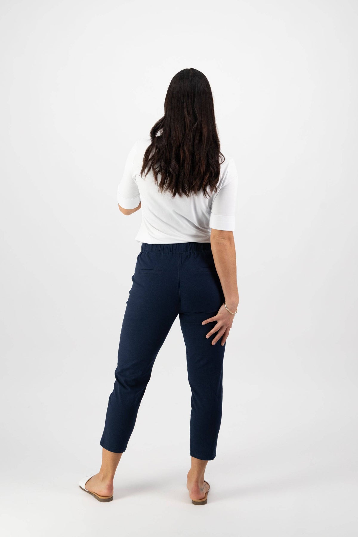 A woman with long dark hair, seen from behind, wears a white short-sleeve top and Vassalli Denver 7/8 Pant - Ink with an elastic waistband, plus sandals. She stands against a plain white background, one hand on her hip.