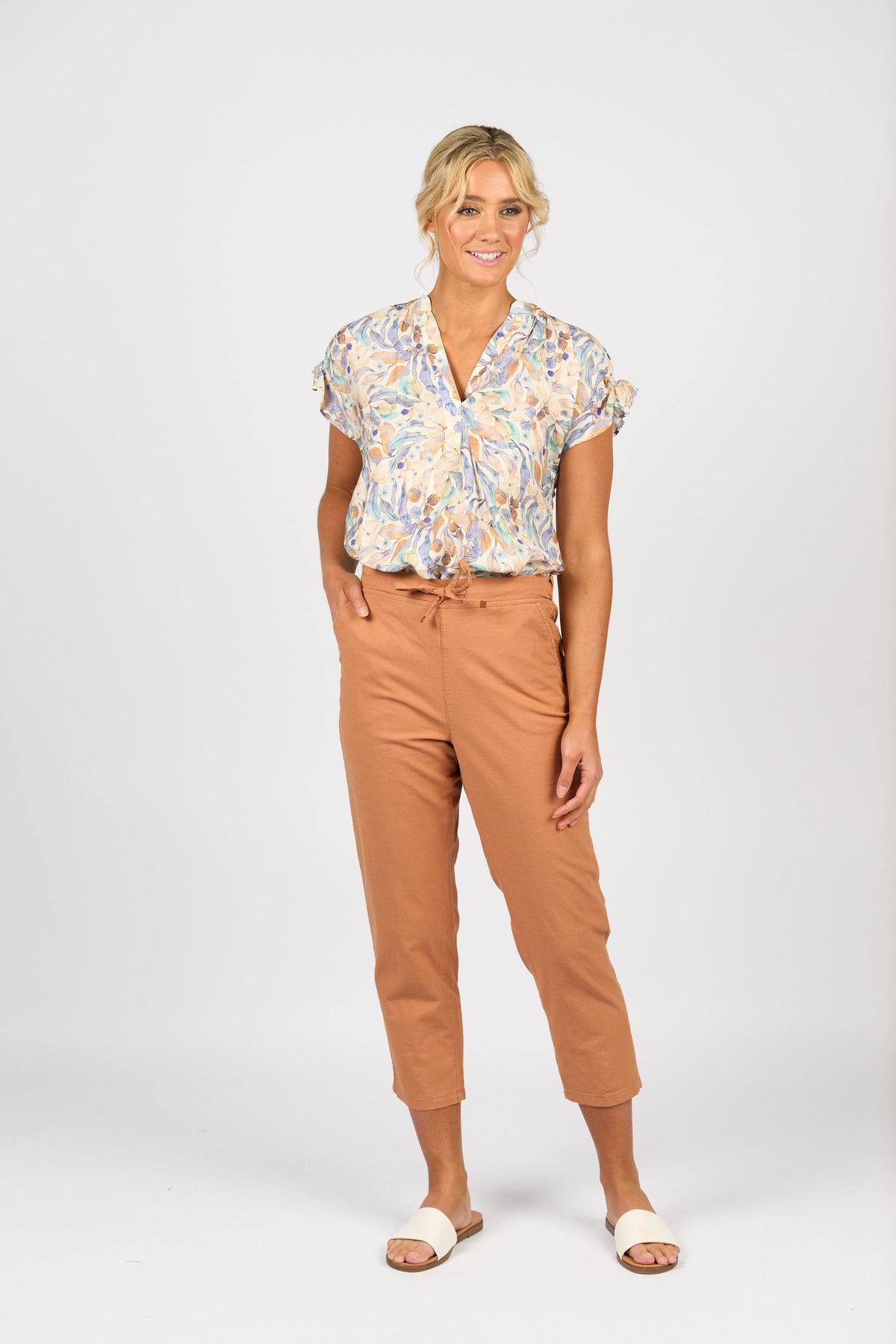 A woman with blonde hair in a loose updo smiles against a plain background, wearing the Vassalli Denver 7/8 Pant in Spice, a patterned short-sleeve cotton blend blouse, and white slide sandals.
