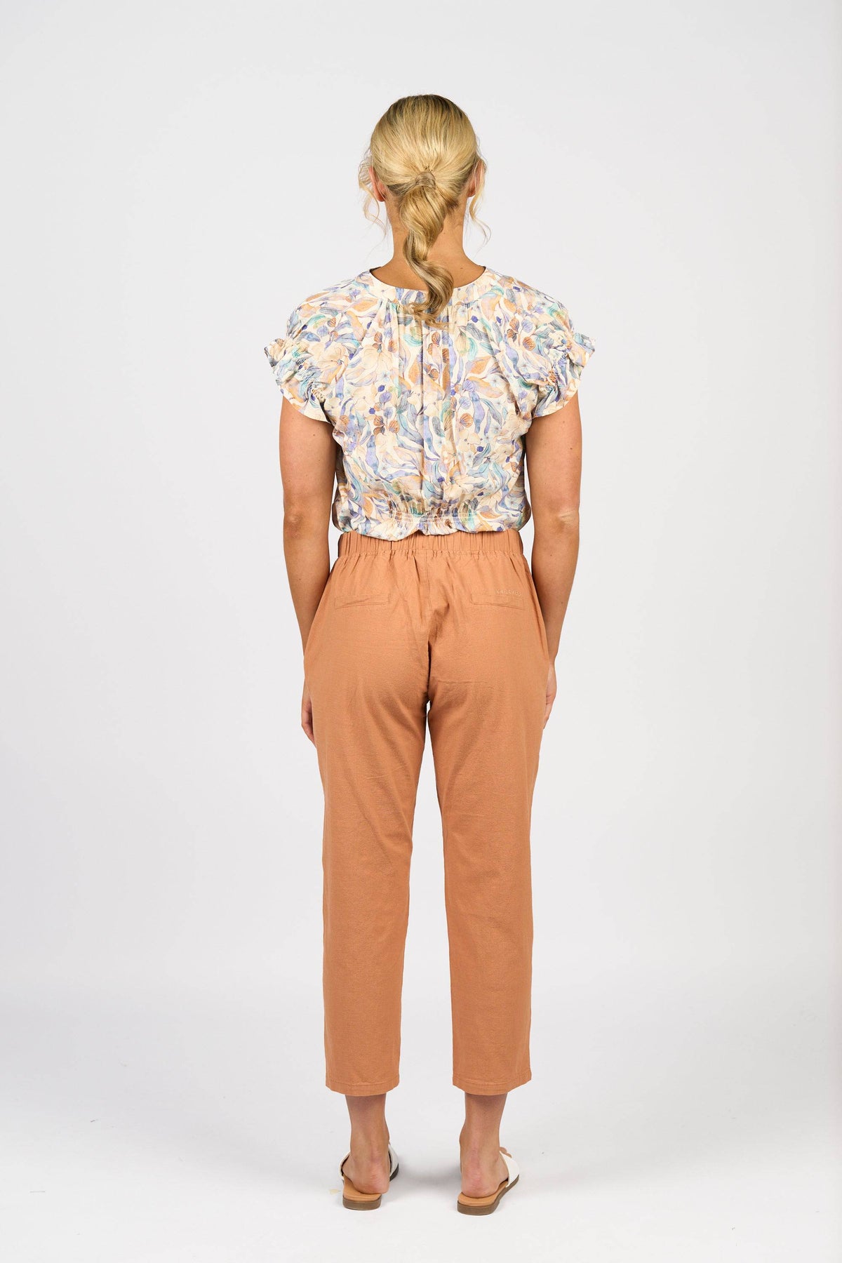 A woman faces away from the camera, wearing Vassalli's Denver 7/8 Pant in Spice with an elastic waistband, a short-sleeve floral blouse, and sandals against a plain white background.
