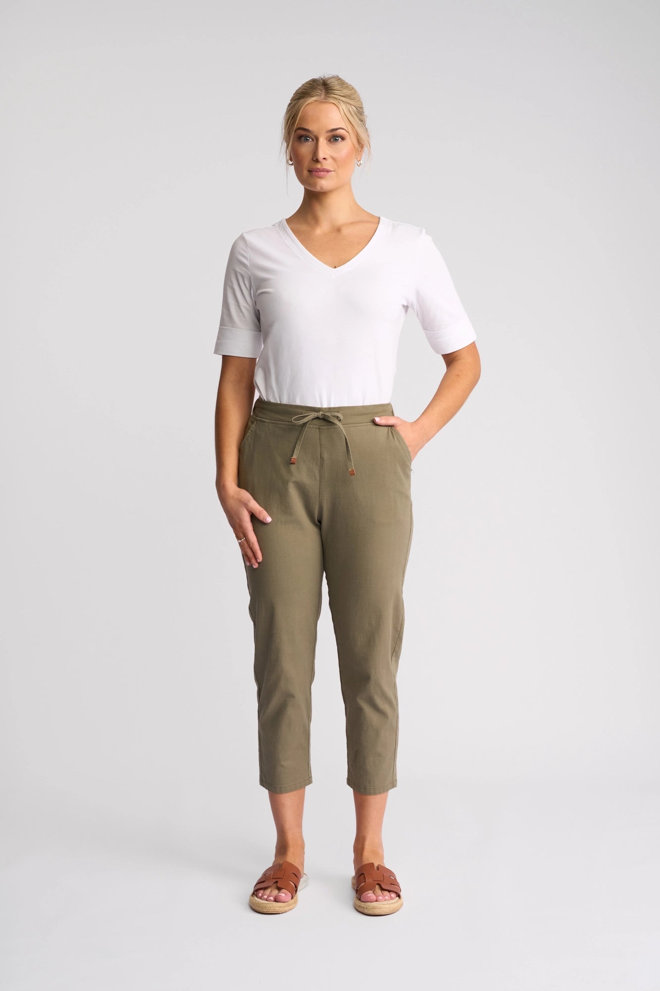 A woman stands facing forward, wearing a white V-neck T-shirt, the Vassalli Denver 7/8 Pant - Army in olive green crafted from stretch cotton with a skinny-leg silhouette, and brown sandals. One hand is in her pocket and the background is plain and light-colored.