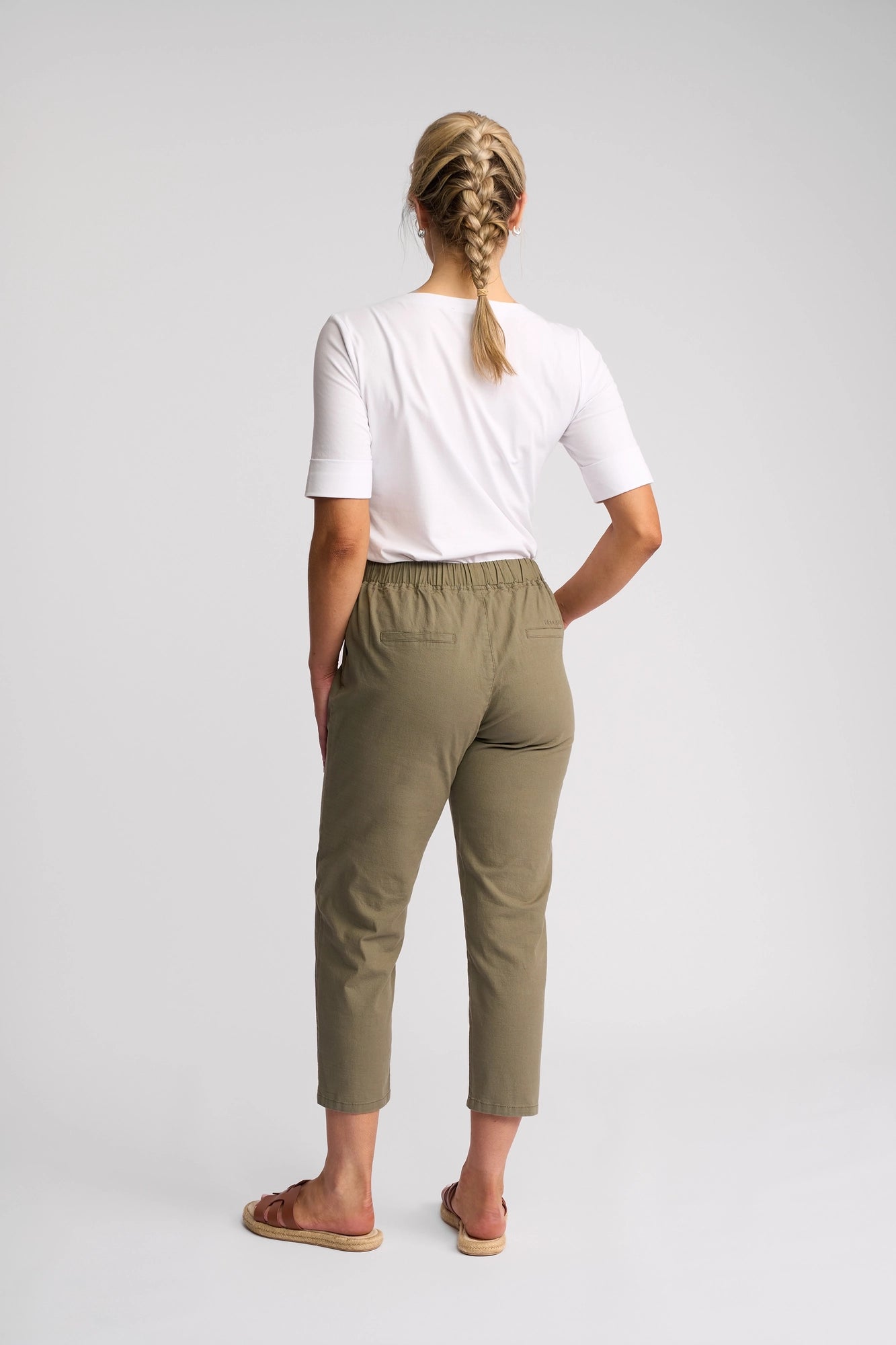 A person with blonde hair in a braid, wearing a white shirt, Vassalli Denver 7/8 Pant - Army with a skinny-leg fit, and sandals, stands facing away against a plain light background.