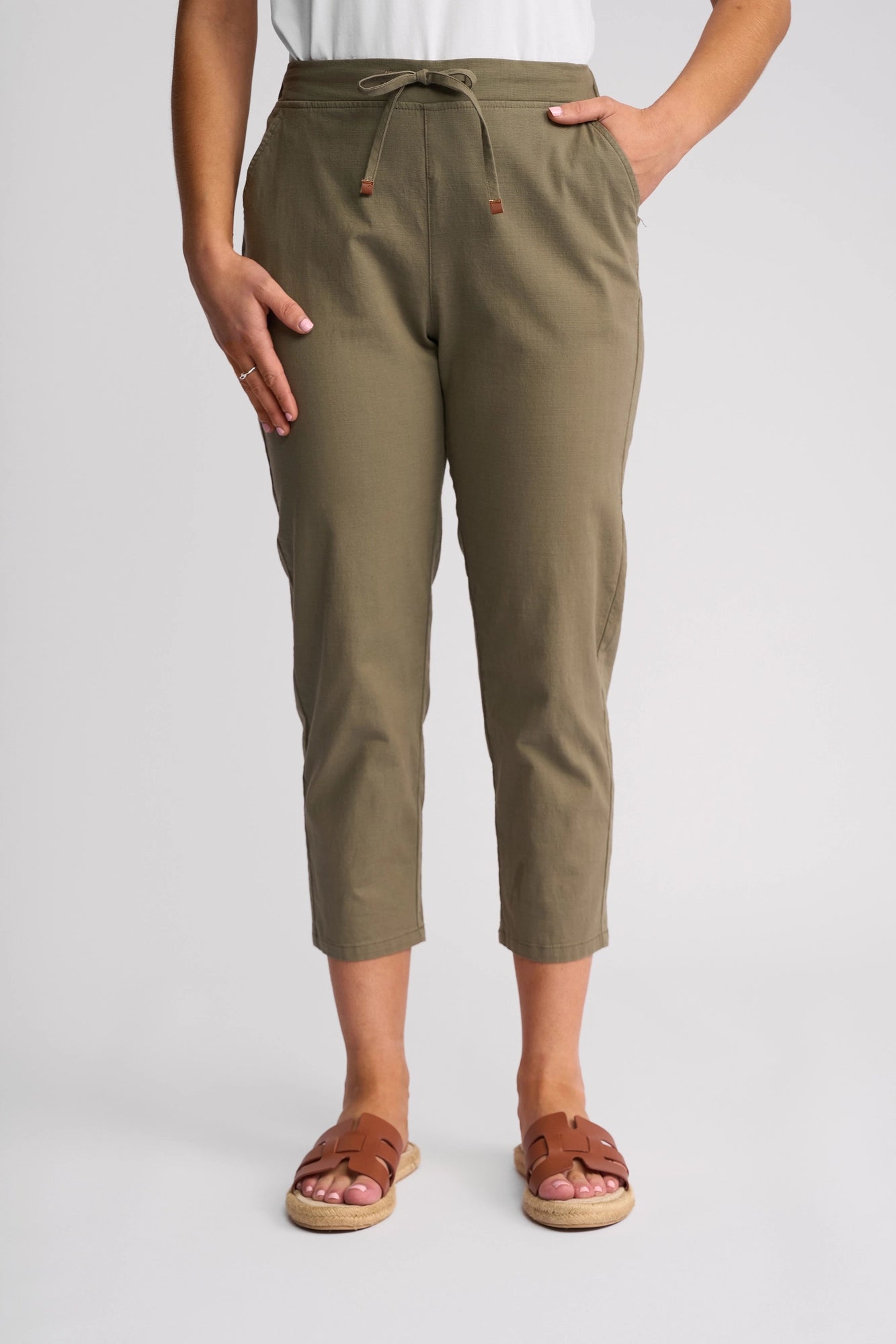 A person stands against a plain light background wearing the Vassalli Denver 7/8 Pant - Army, featuring an olive green stretch cotton fabric and drawstring waist, paired with a white top and brown sandals.