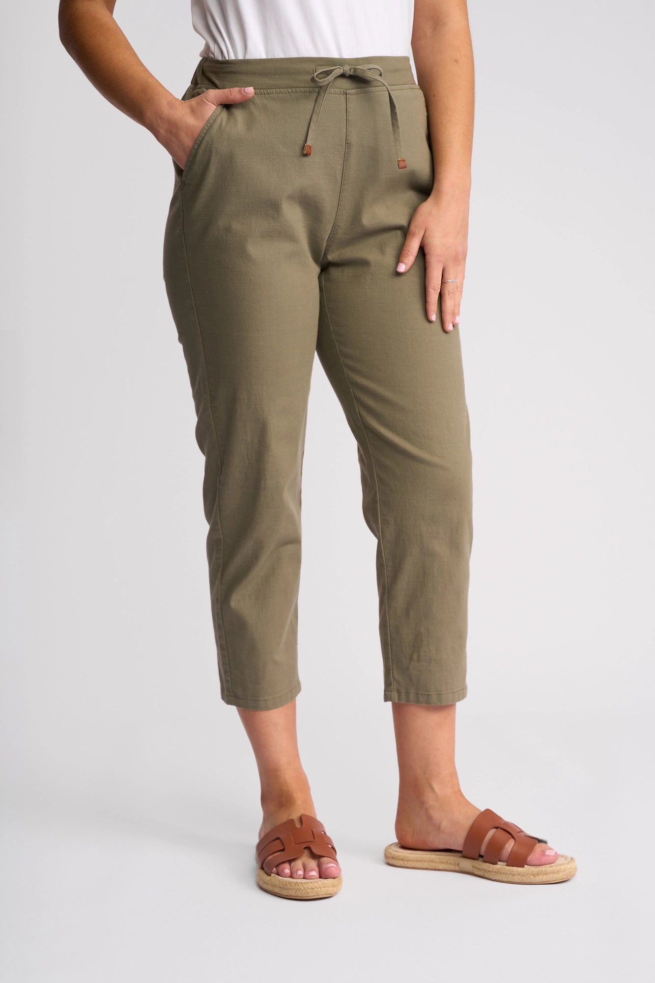 A person wearing Vassalli’s Denver 7/8 Pant in Army, paired with a white top and brown sandals, stands against a plain white background with one hand in their pocket and the other resting on their thigh.