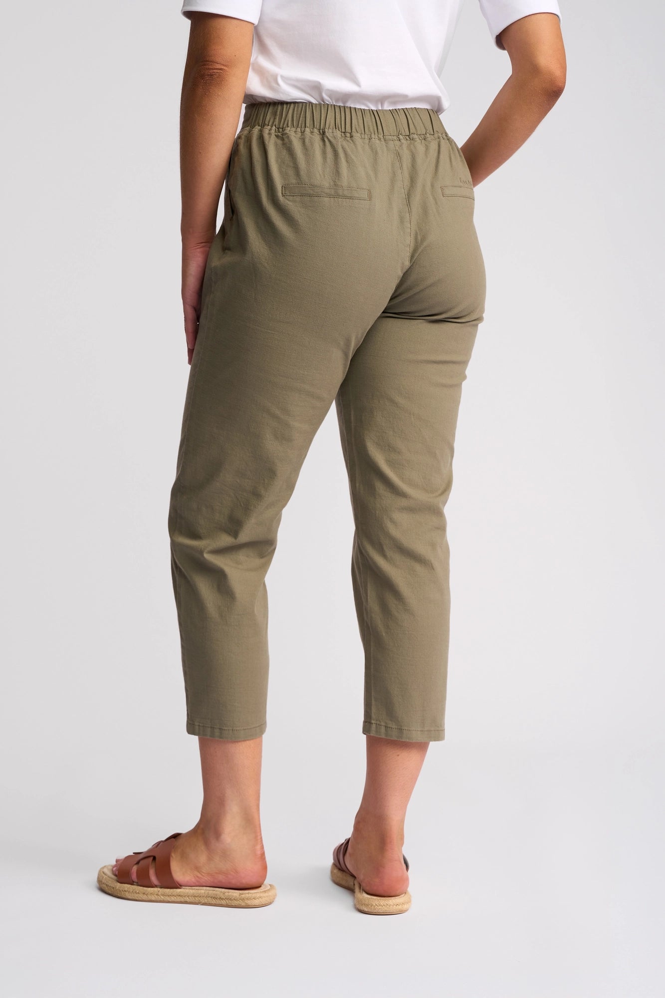 A person stands facing away, wearing Vassalli’s Denver 7/8 Pant - Army in a stretch cotton, skinny-leg style. Paired with a white shirt and brown sandals, only the lower back and legs are visible against a white background.