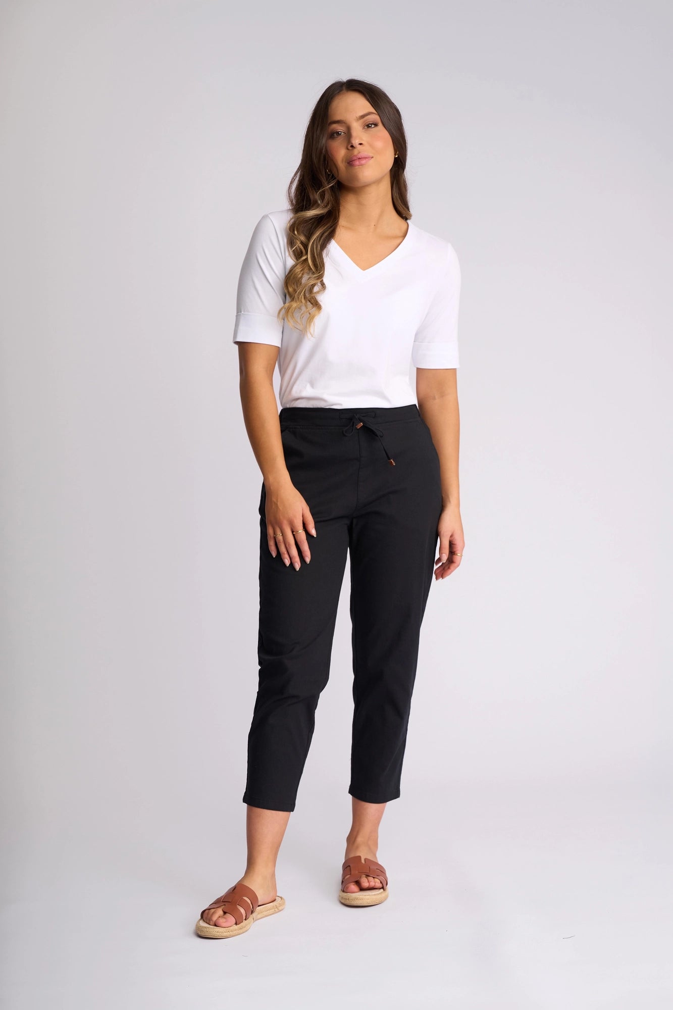 A woman stands against a plain background, wearing a white V-neck T-shirt paired with the Vassalli Denver 7/8 Pant in black. The skinny-leg design, crafted from stretch cotton, perfectly complements her brown sandals. She has long, wavy hair and a relaxed expression.