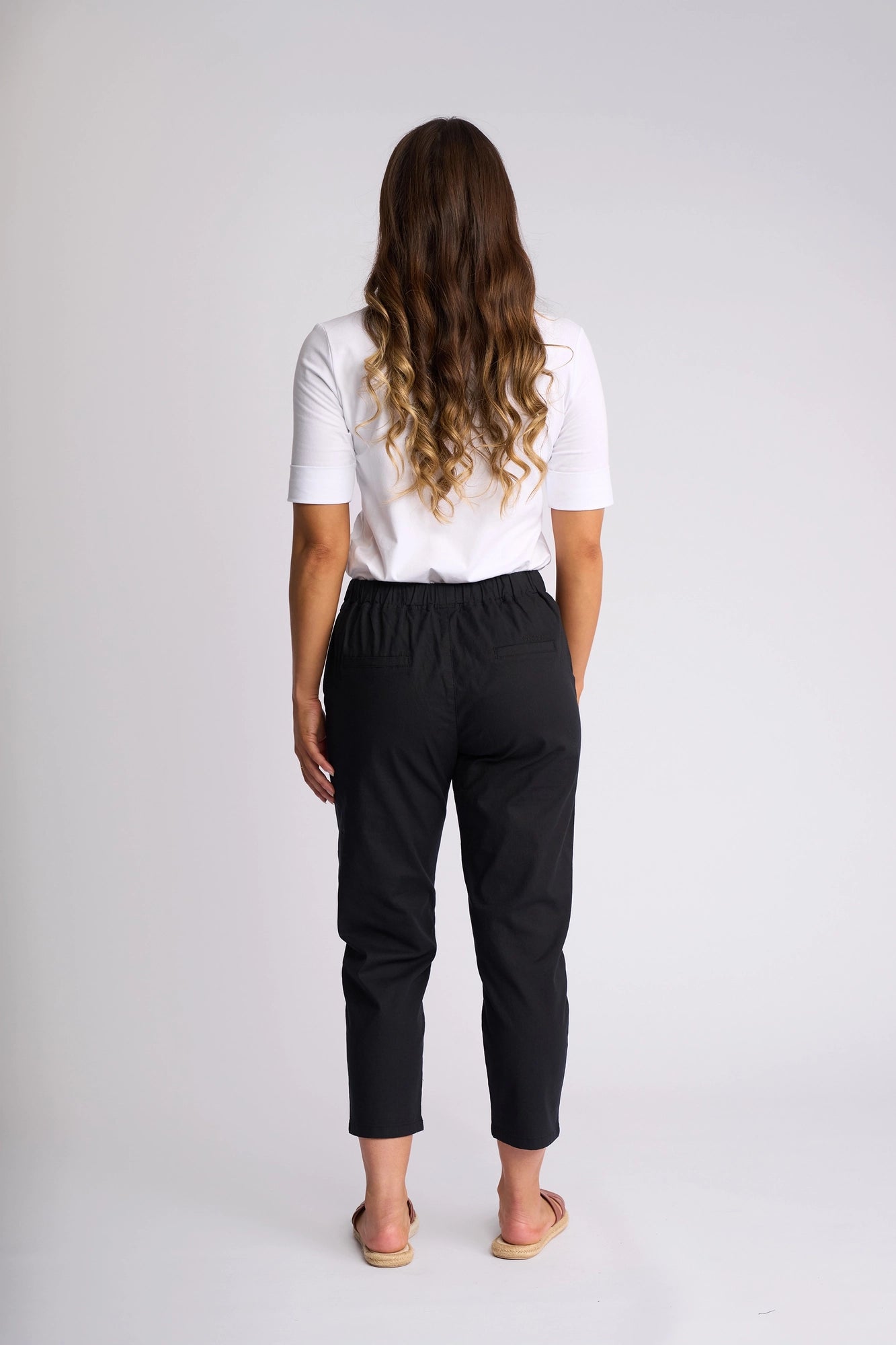 A woman with long wavy hair, wearing a white short-sleeve top and Vassalli Denver 7/8 Pant in black, paired with sandals, stands facing away from the camera against a plain light background.