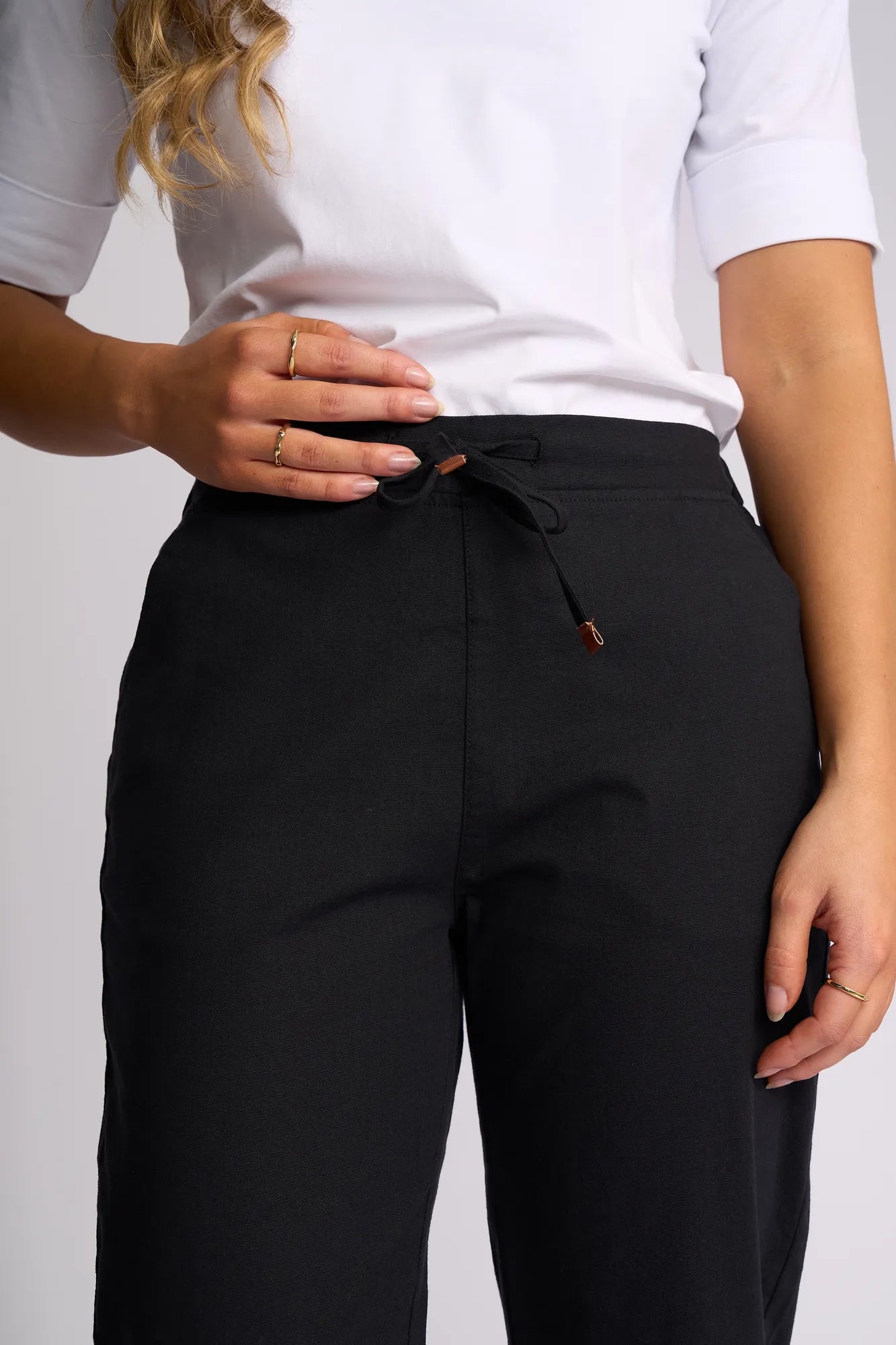 A person wearing a white shirt and Vassalli’s Denver 7/8 Pant in black stands with one hand resting on the waistband, showcasing gold rings on their fingers. The skinny-leg pant, made from comfortable stretch cotton, brings a sleek and modern vibe to the outfit.