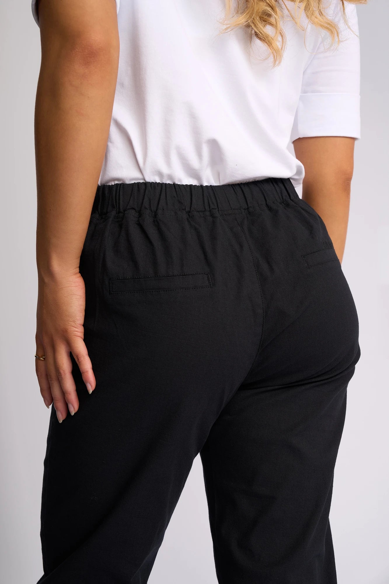 A person is shown from the back wearing a white shirt and Vassalli’s Denver 7/8 Pant in black, with their hand resting on their hip. The image focuses on the lower back and upper legs, emphasizing the flattering skinny-leg silhouette and comfortable stretch cotton fabric of these pants.