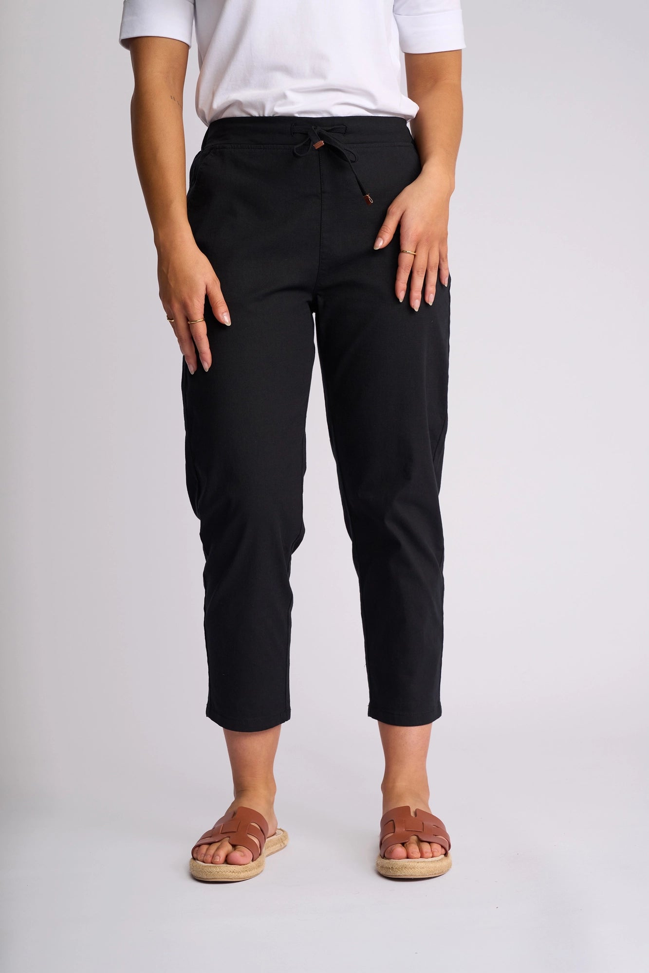A person wearing a white shirt, the Vassalli Denver 7/8 Pant in Black, and brown sandals stands against a plain light background. Only the lower half of the body is visible.