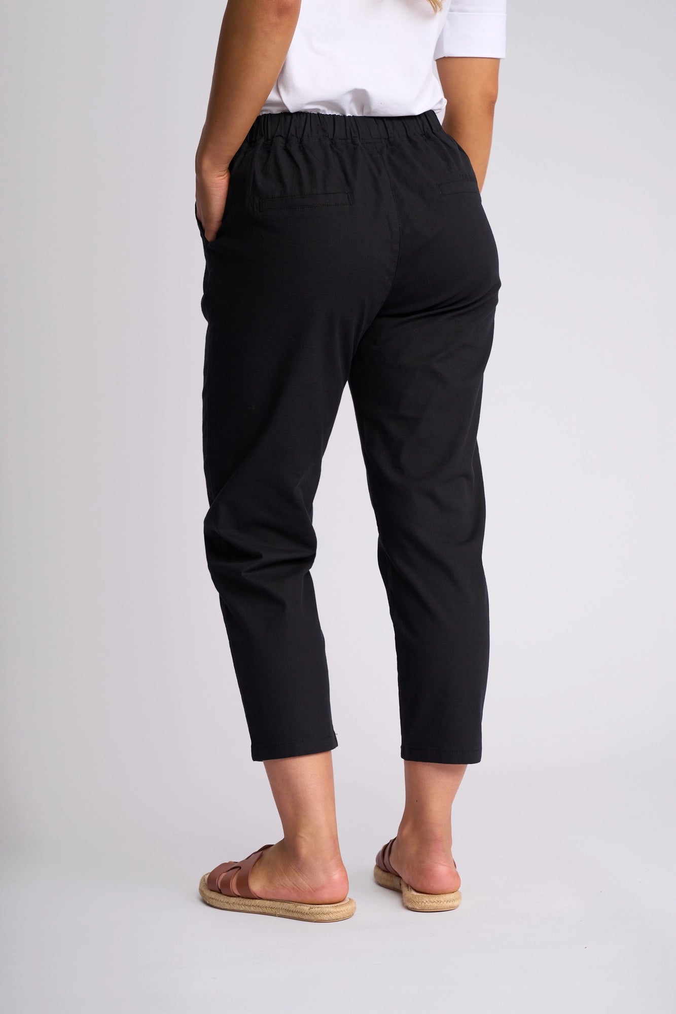 A person wearing the Vassalli Denver 7/8 Pant in Black, paired with a white top and brown sandals, stands with hands in pockets facing away from the camera against a plain light background. The skinny-leg pant features comfortable stretch cotton for a sleek yet relaxed appearance.
