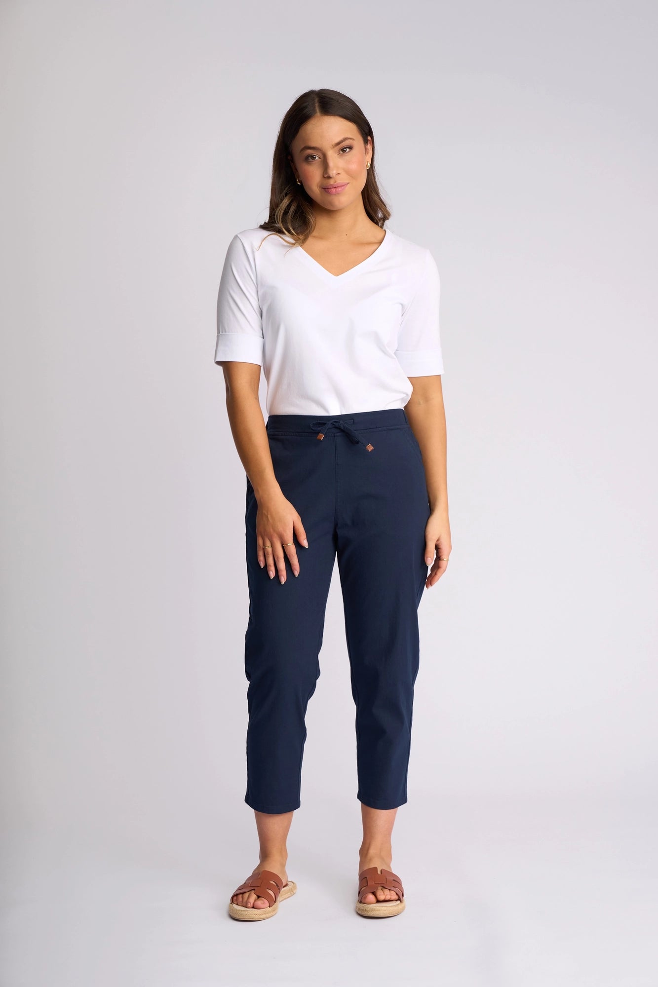 A woman stands against a plain background wearing a white V-neck shirt, the Vassalli Denver 7/8 Pant in Ink featuring a tie waist and comfortable stretch cotton fabric, and brown sandals. Her hair is down and she looks at the camera with a slight smile.
