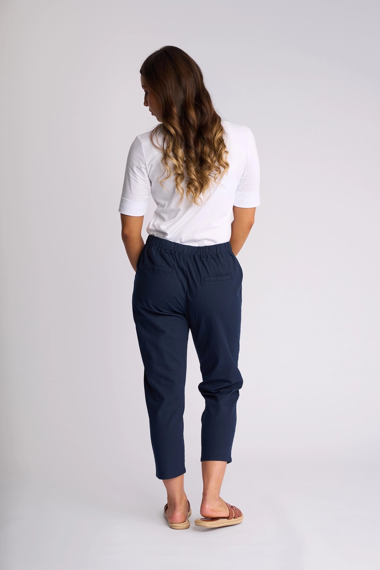Woman with long wavy hair stands facing away, wearing a white shirt, Vassalli Denver 7/8 Pant in Ink with a stretch cotton skinny-leg silhouette, and sandals against a plain light background.