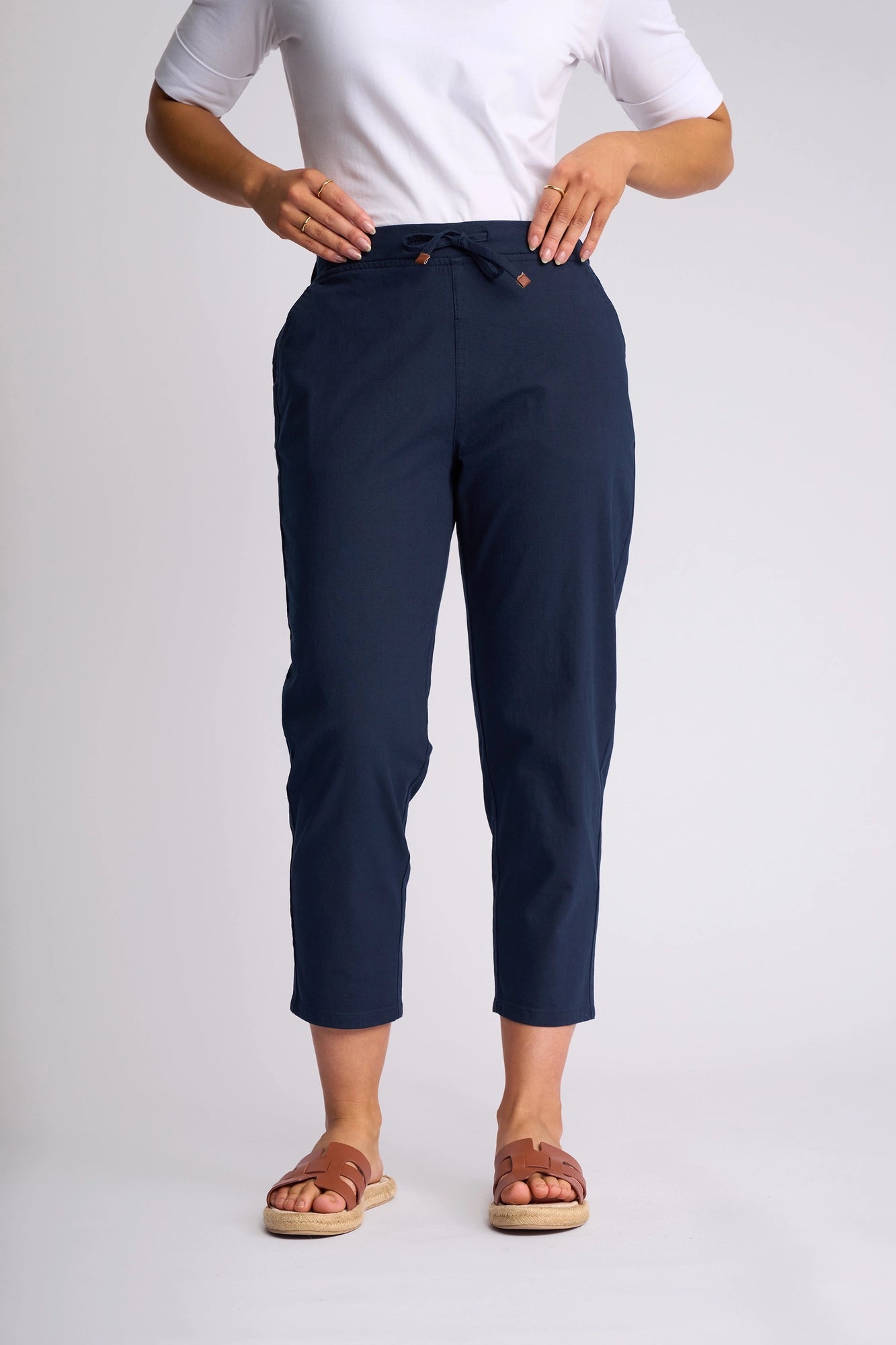 A person wearing a white shirt and Vassalli Denver 7/8 Pant in Ink, featuring an ankle-length, skinny-leg fit with a comfortable drawstring waist and crafted from stretch cotton, paired with brown sandals and standing against a plain light background.