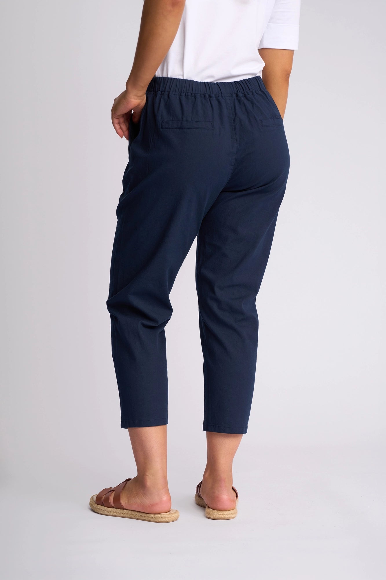 A person wearing a white short-sleeve top, brown sandals, and Vassalli’s Denver 7/8 Pant in Ink—a skinny-leg style crafted from stretch cotton—stands against a plain light background, shown from the back.