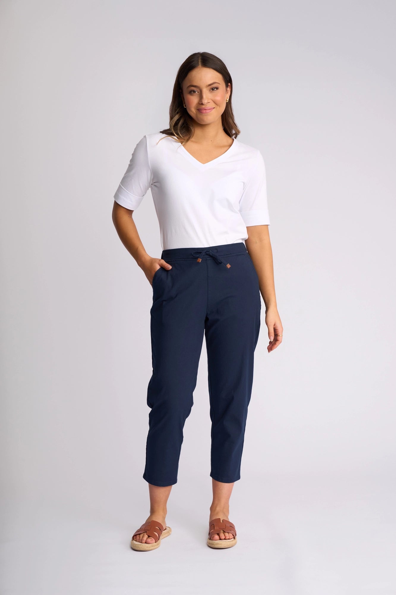 A woman stands against a plain background wearing a white short-sleeve V-neck top, the Vassalli Denver 7/8 Pant in Ink featuring a skinny-leg silhouette crafted from stretch cotton, and brown sandals, with one hand in her pocket and a relaxed expression.