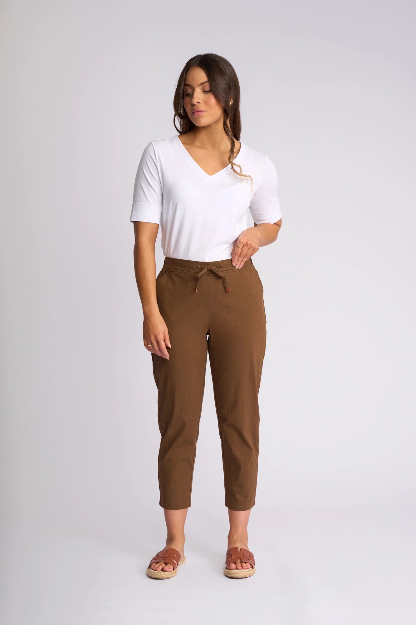 A woman stands against a light background, wearing a white V-neck top, brown sandals, and the Vassalli Denver 7/8 Pant in Pecan—crafted from stretch cotton with a drawstring waist. She has long hair and looks slightly to the side.