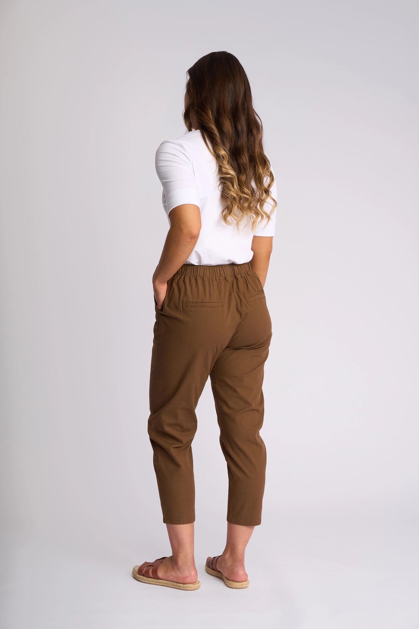 A woman with long wavy hair, wearing a white top and the Vassalli Denver 7/8 Pant in Pecan—a stylish stretch cotton skinny-leg pant—paired with sandals, stands facing away with her hands in her pockets against a plain light background.